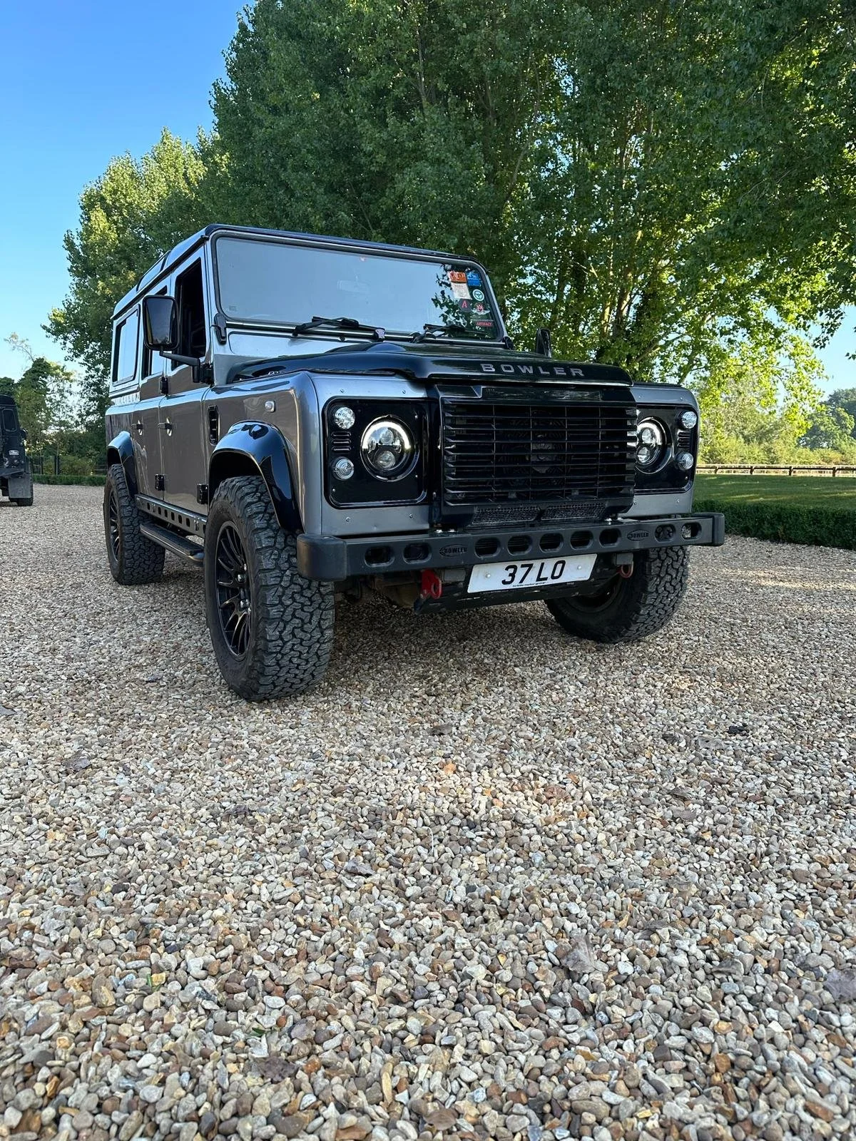 A black and gray off-road Land Rover Defender parked on a gravel surface under a tree with green foliage in the background.