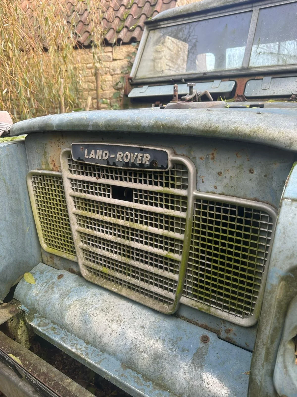 Close-up of an old, rusty Land Rover vehicle front, showing the grille and badge, with moss and dirt.