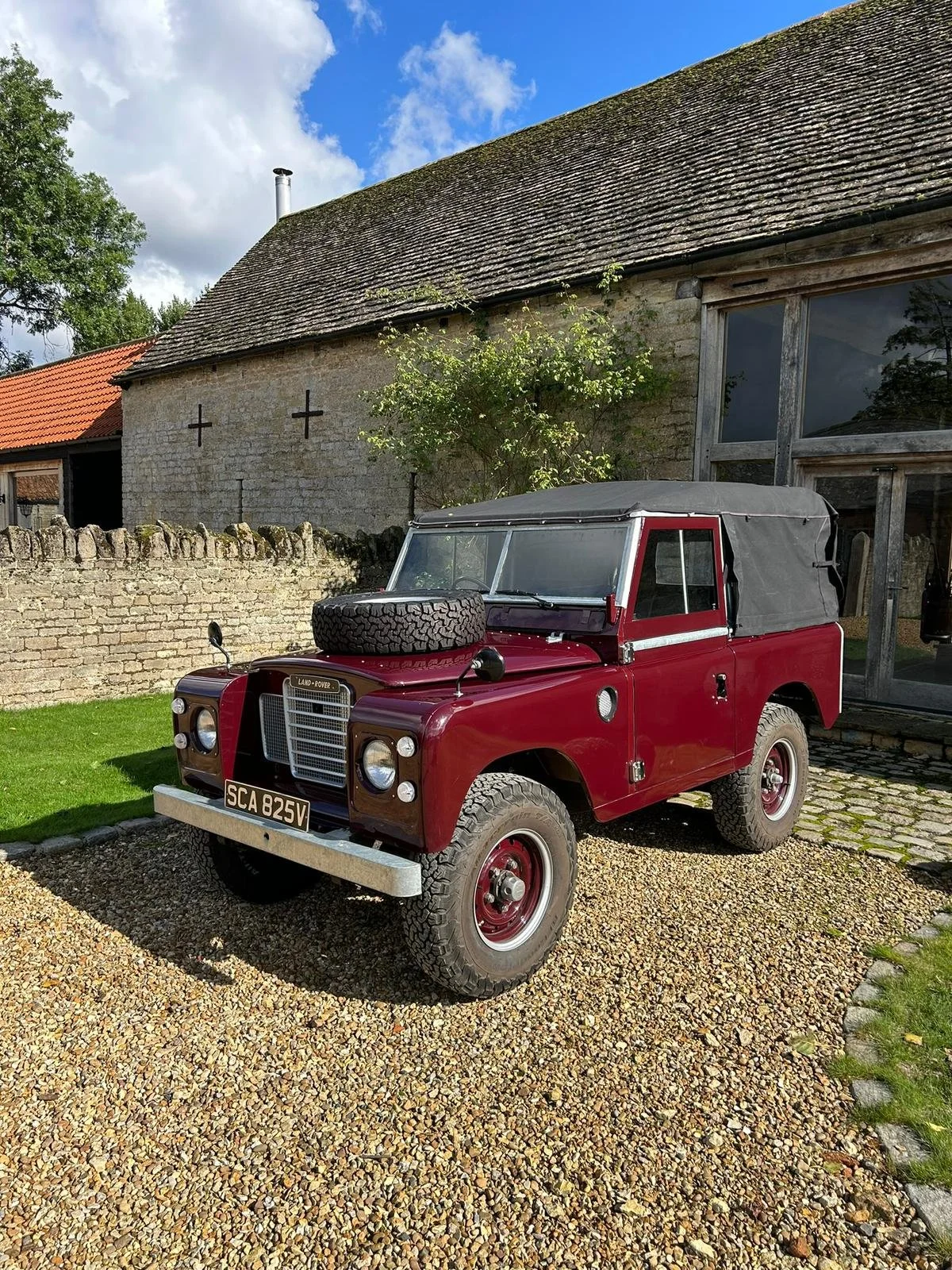 Red vintage Land Rover with a black soft top and a spare tire on the hood, parked on a gravel driveway in front of a rustic stone house with a shingled roof.