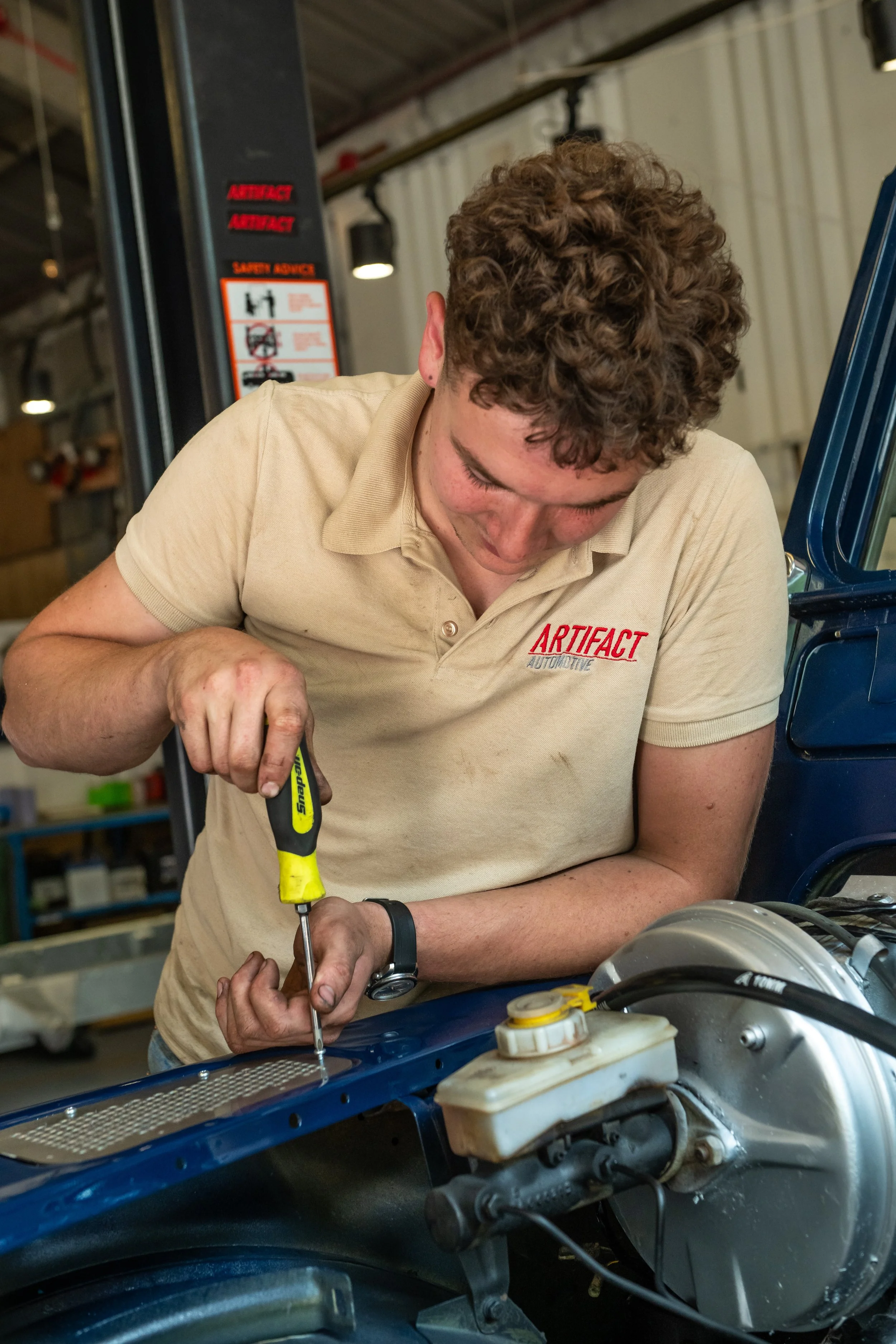 A mechanic working on a vehicle's engine compartment using a screwdriver in a garage or workshop.