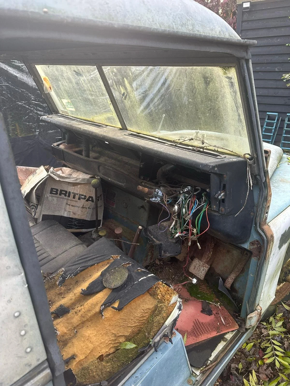 Inside of an old, abandoned vehicle with a rusted dashboard, exposed wires, torn seat, and moss growing on the floor.