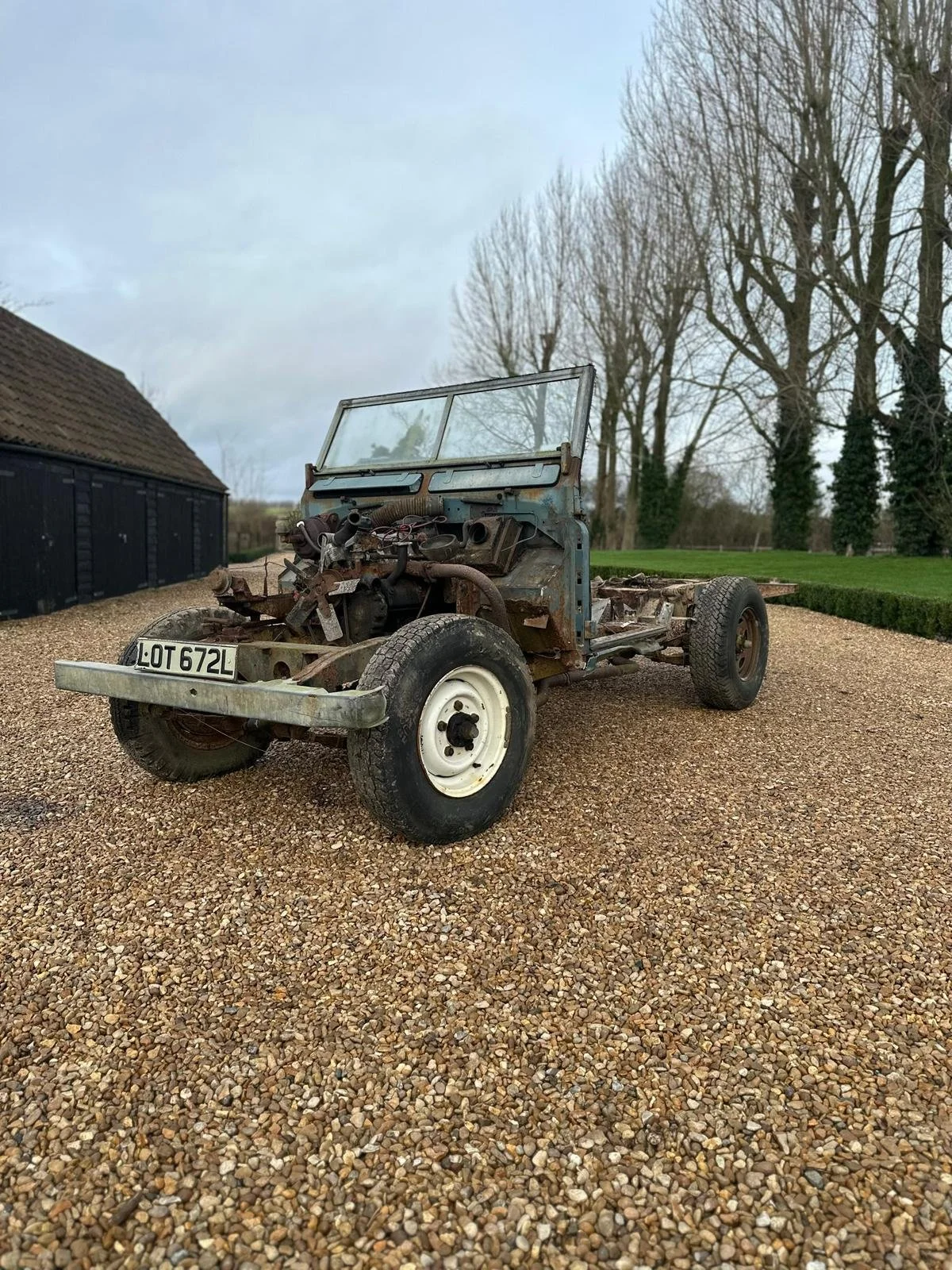 Old, rusted, disassembled vehicle with missing parts and no engine, parked on a gravel driveway with trees and a shed in the background.