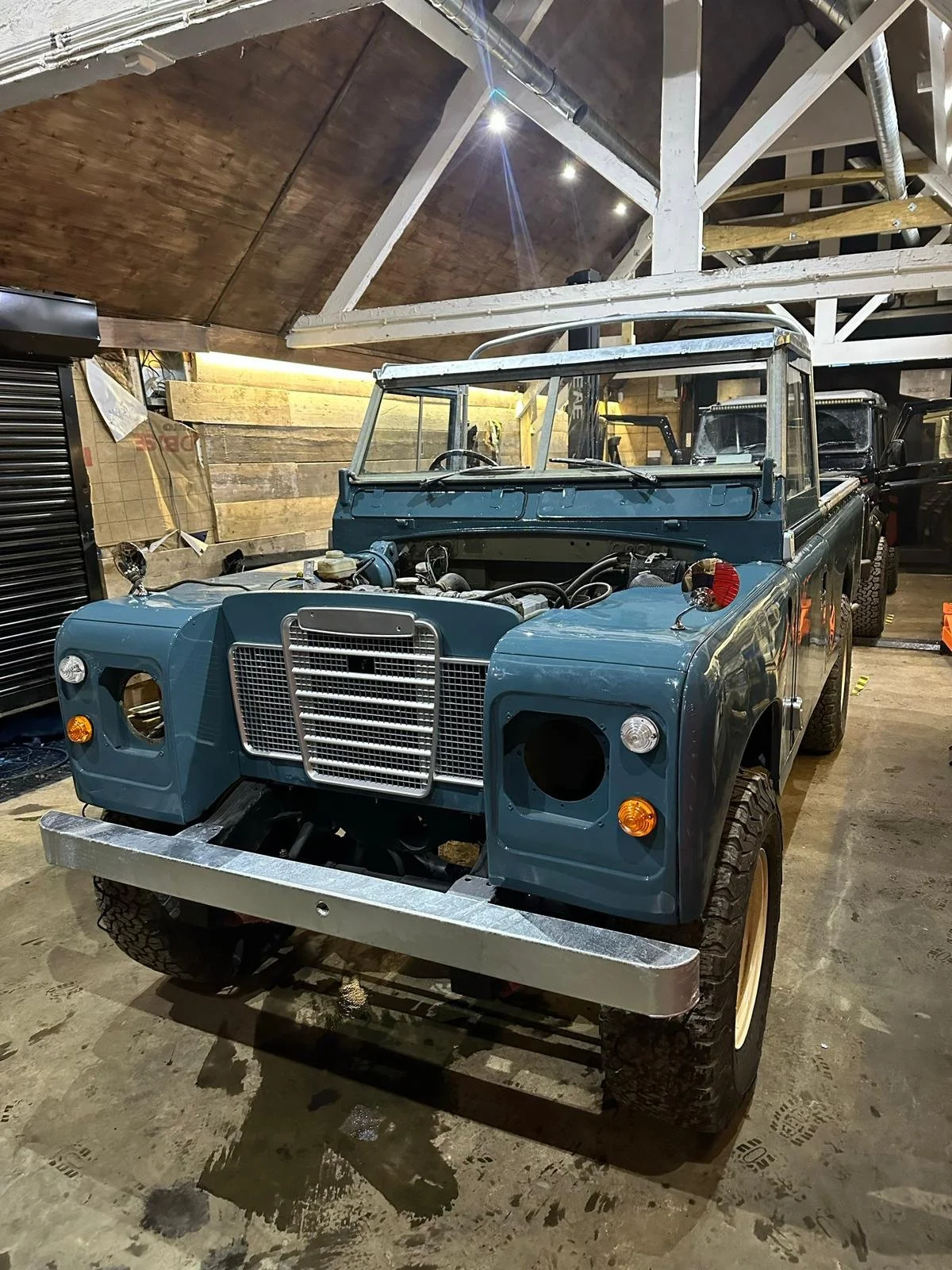 A vintage blue Land Rover vehicle inside a garage, with some parts missing, showing the engine bay and front grille. The garage has a wooden ceiling and various tools and equipment around.