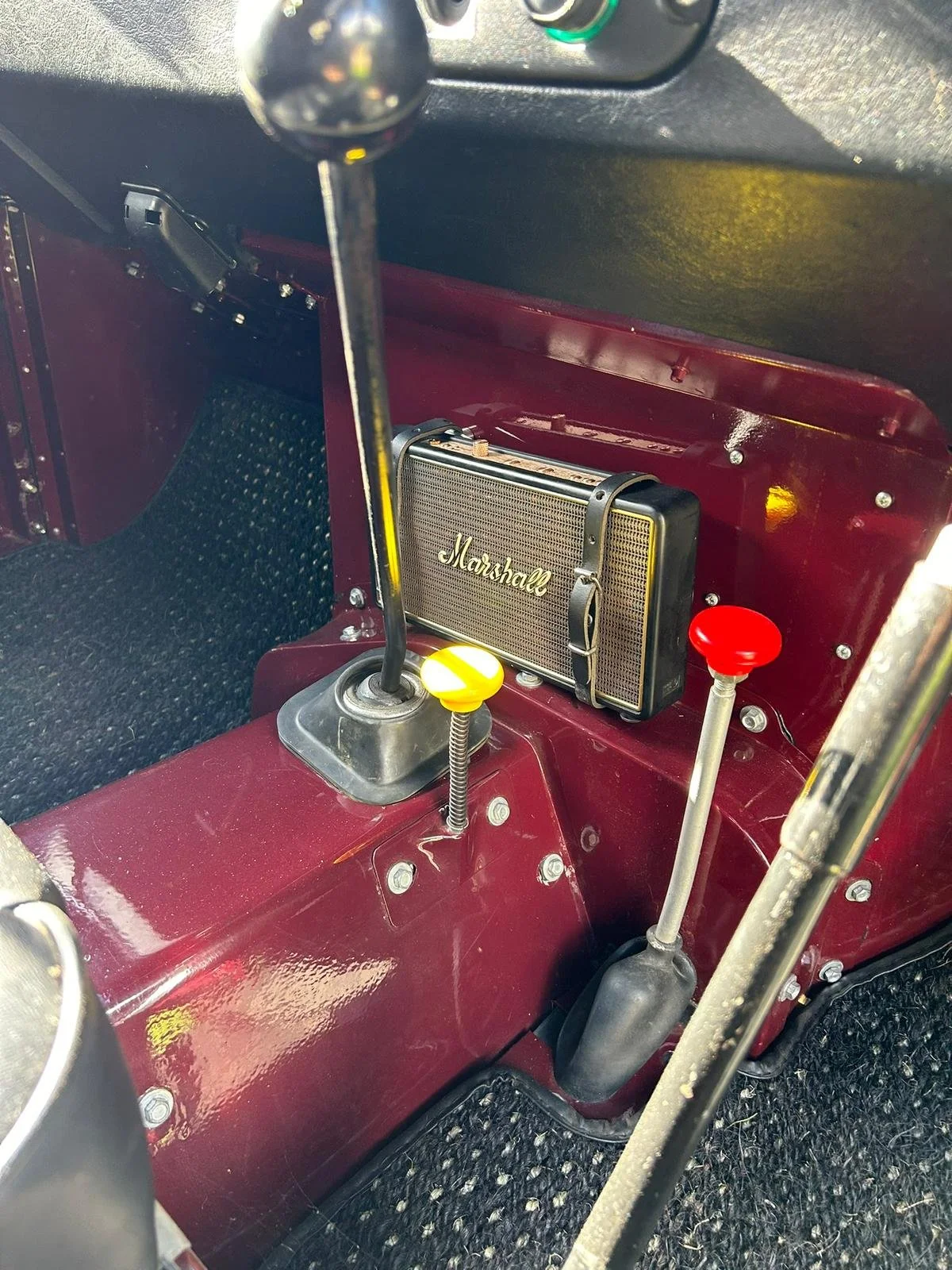 Close-up of the interior of a vintage car dashboard showing a Marshall speaker, gear shift with a yellow button, and two control levers with red and yellow knobs.