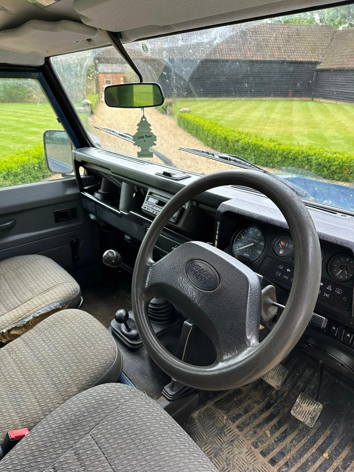 Interior view of an older Land Rover vehicle with a manual transmission, showing steering wheel, gauges, vintage seats, and a view through the windshield of a gravel driveway, green lawn, and a wooden shed.