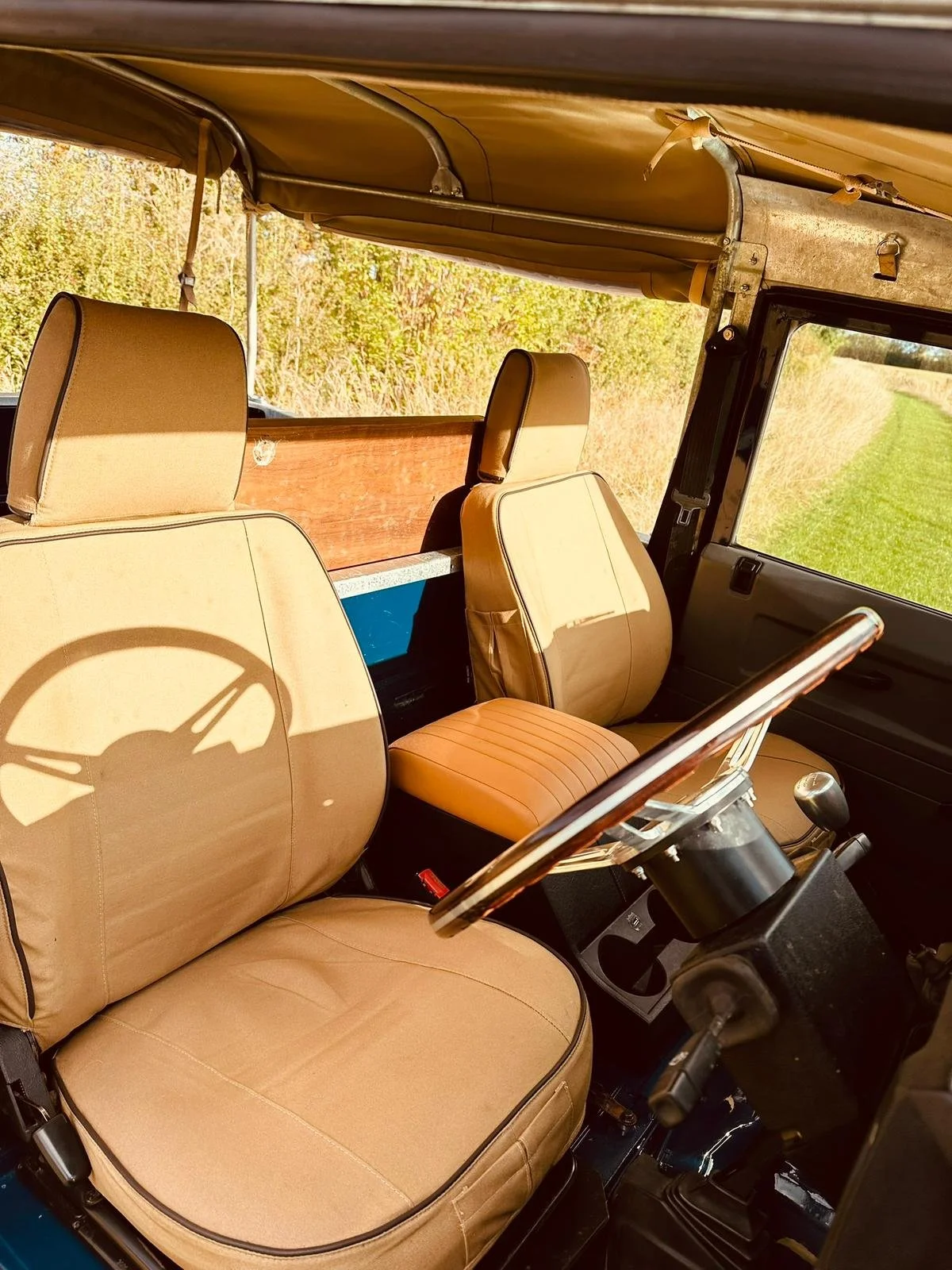 Interior of a vintage vehicle with tan seats, black steering wheel, and a gear shift, parked on a grassy and wooded area.