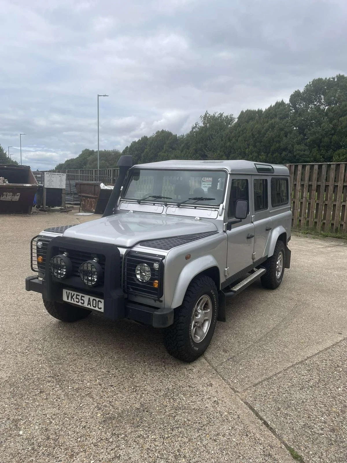 Silver Land Rover Defender parked on a gravel lot with a wooden fence and trees in the background under a cloudy sky.