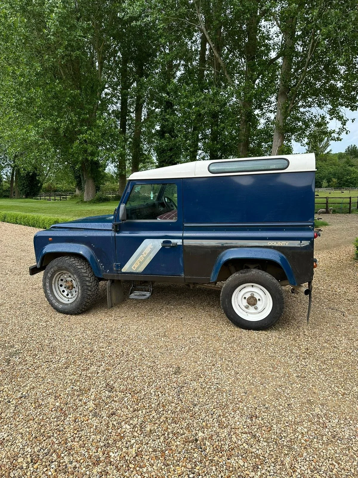 A dark blue vintage utility vehicle with a white top, parked on a gravel surface with green trees in the background.