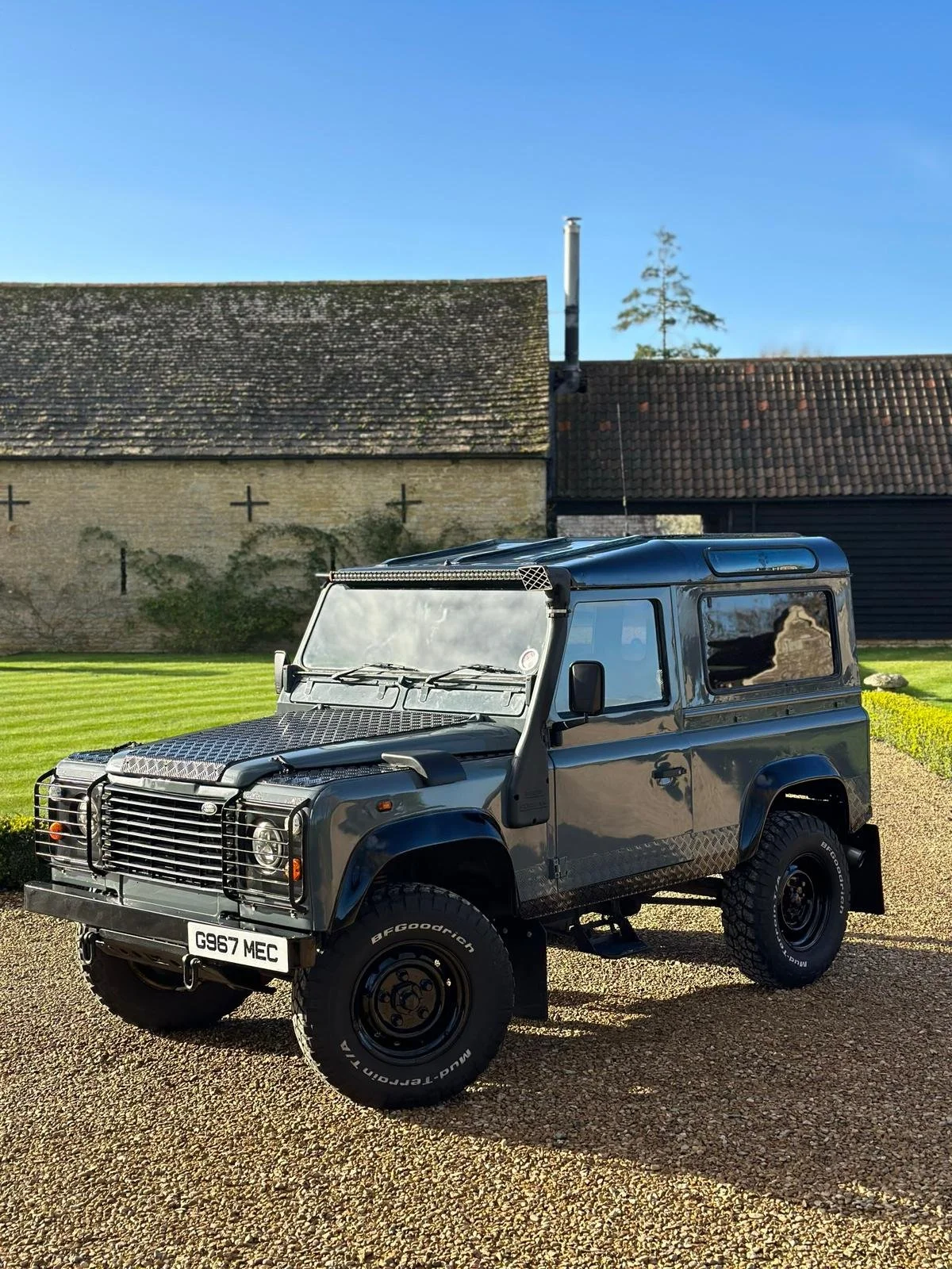 A black, custom-built off-road vehicle with large tires, metal accents, and a roof rack parked on a gravel driveway in front of a grassy area with a stone building and trees in the background.