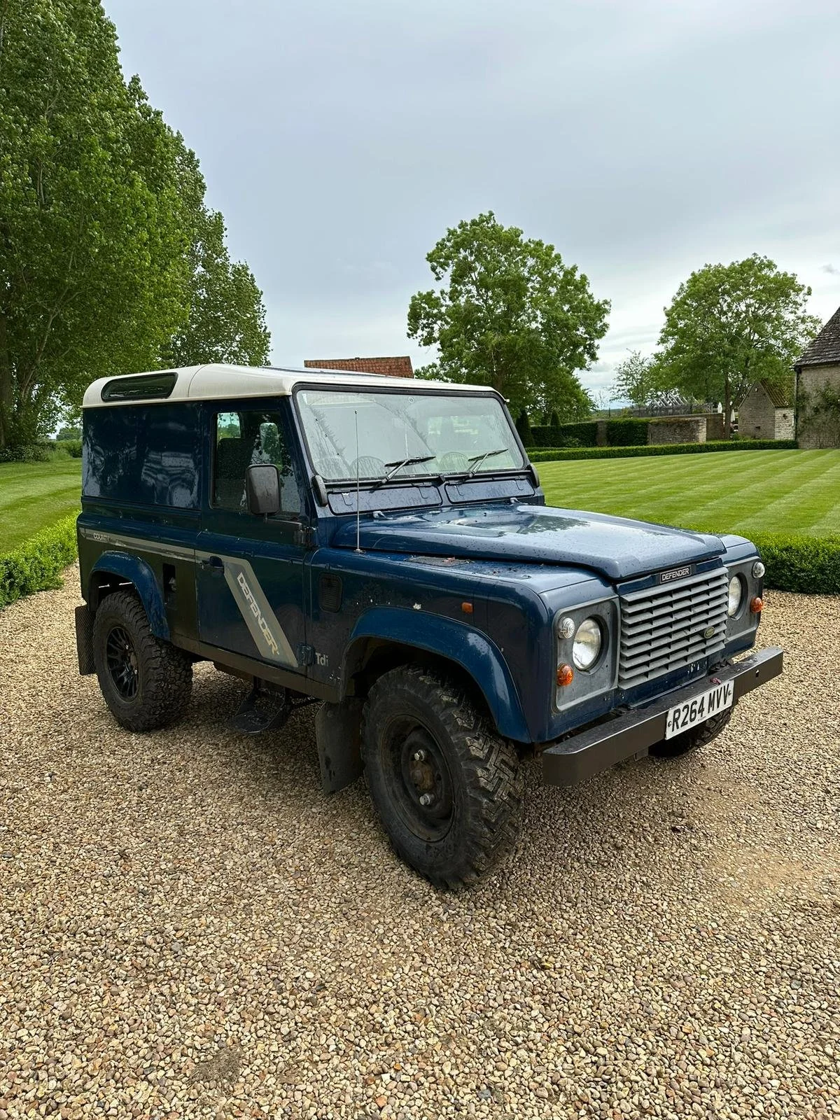 A blue Land Rover Defender vehicle parked on a gravel driveway with grass and trees in the background.