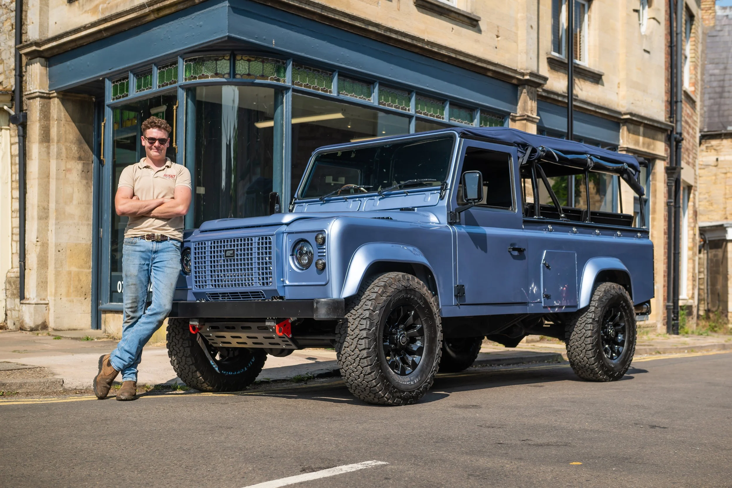 A man wearing sunglasses, a beige T-shirt, jeans, and boots leaning against a blue Land Rover Defender parked on the street in front of a building with large glass windows.