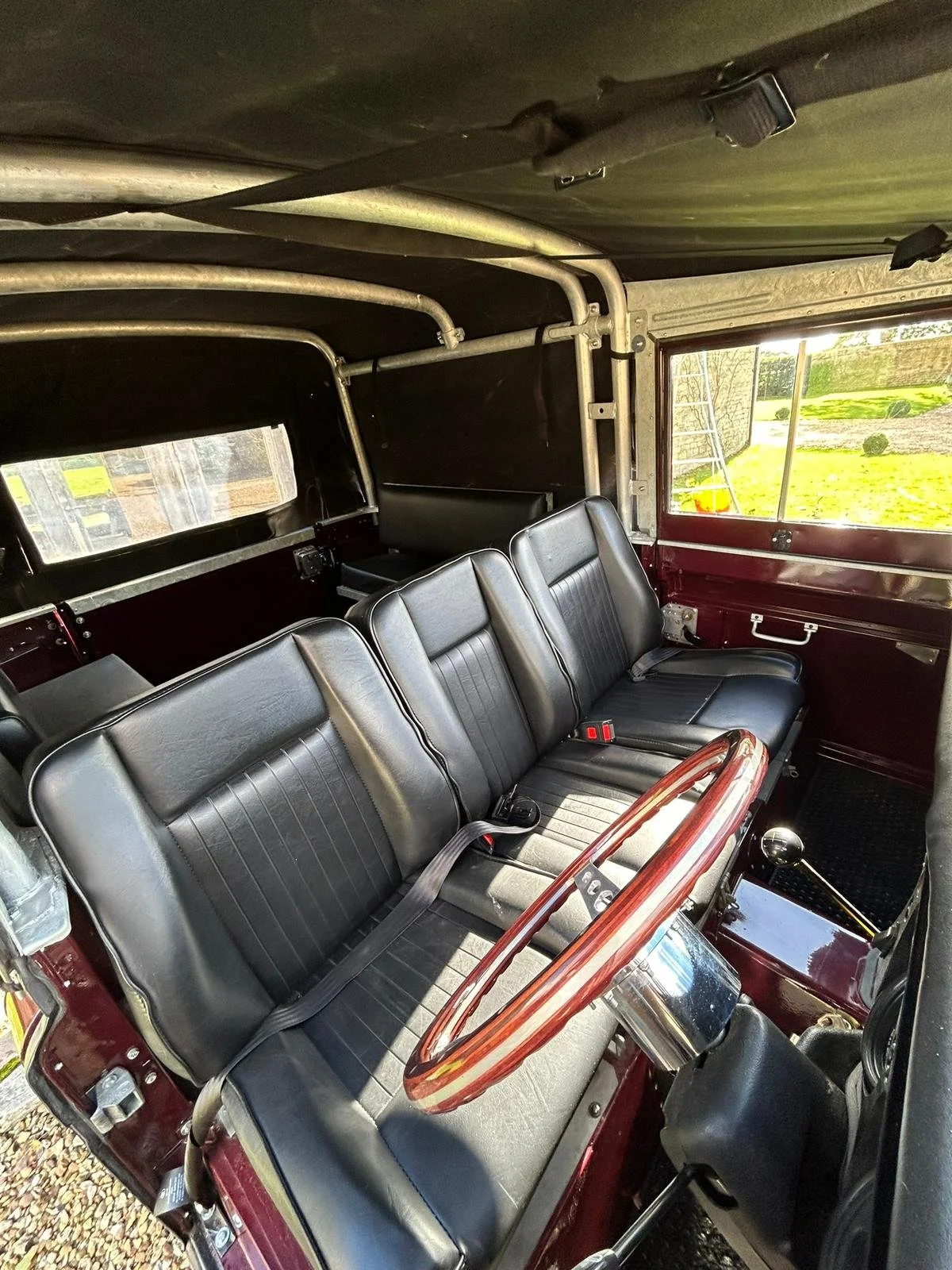 Interior of a vintage vehicle with black leather seats, a wooden steering wheel, and a gear shift, with windows revealing a grassy outdoor area.