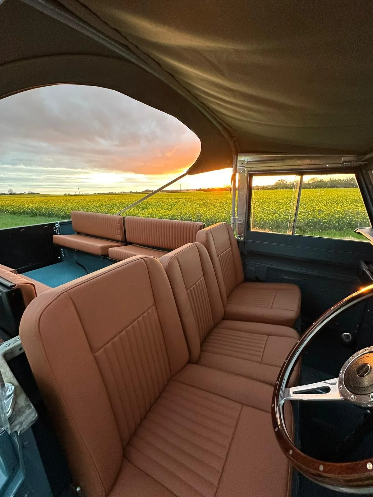 Interior of a vintage vehicle with tan leather seats, large front windshield, and a sliding window showing a sunset over a green field.