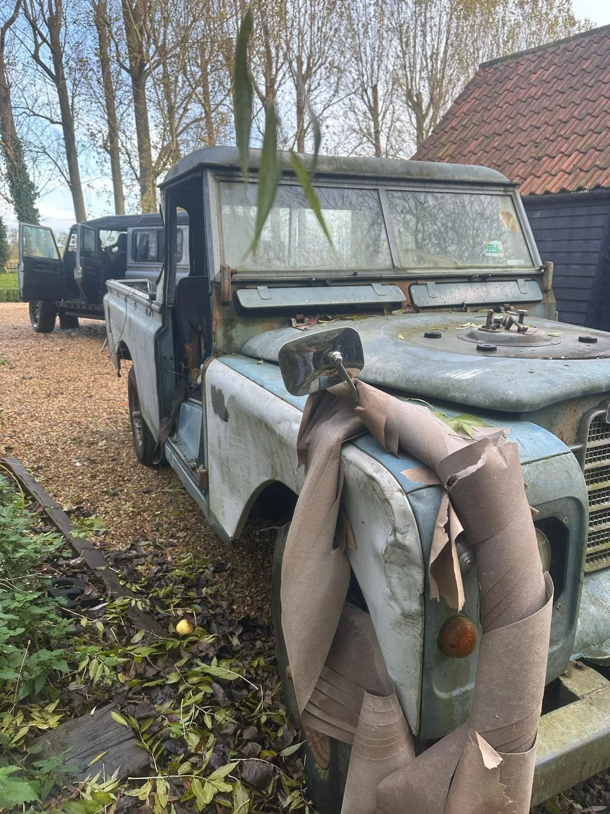 Old, rusted vintage four-wheel drive vehicle with a battered appearance, parked outdoors with other vehicles in the background, surrounded by fallen leaves and foliage.