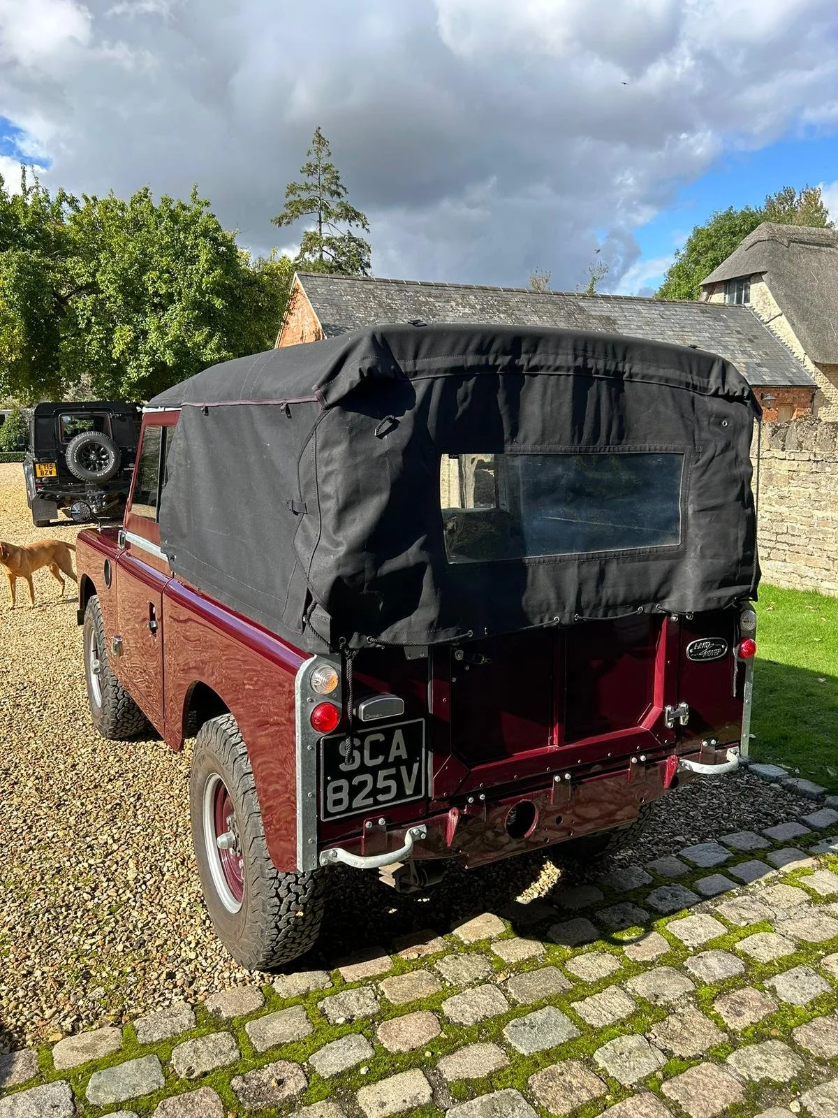 Red Land Rover Series vehicle with a black soft top roof parked on cobblestones in a garden area.