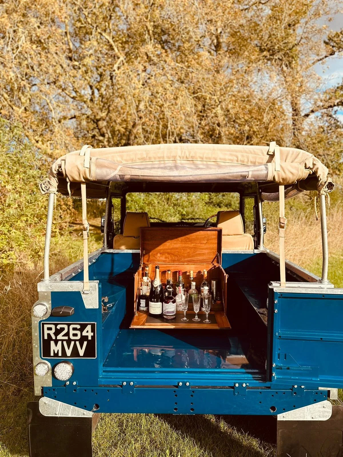 A blue vintage utility vehicle with a wooden bar and bottles of alcohol, glasses, and a wooden box inside, set outdoors with trees in the background.