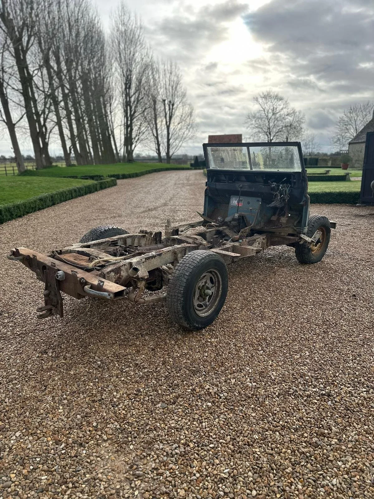 A stripped-down, rusty vehicle frame with no body panels, sitting on a gravel driveway in a rural area with trees, grass, and cloudy sky in the background.