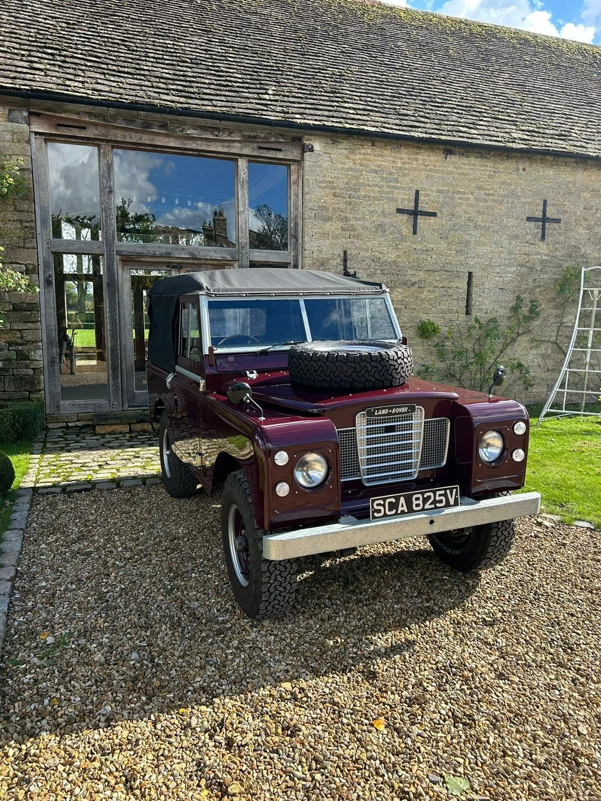 A vintage maroon Land Rover with a spare tire on the hood, parked on a gravel driveway in front of a rustic stone house with large windows and a wooden door, under a partly cloudy sky.
