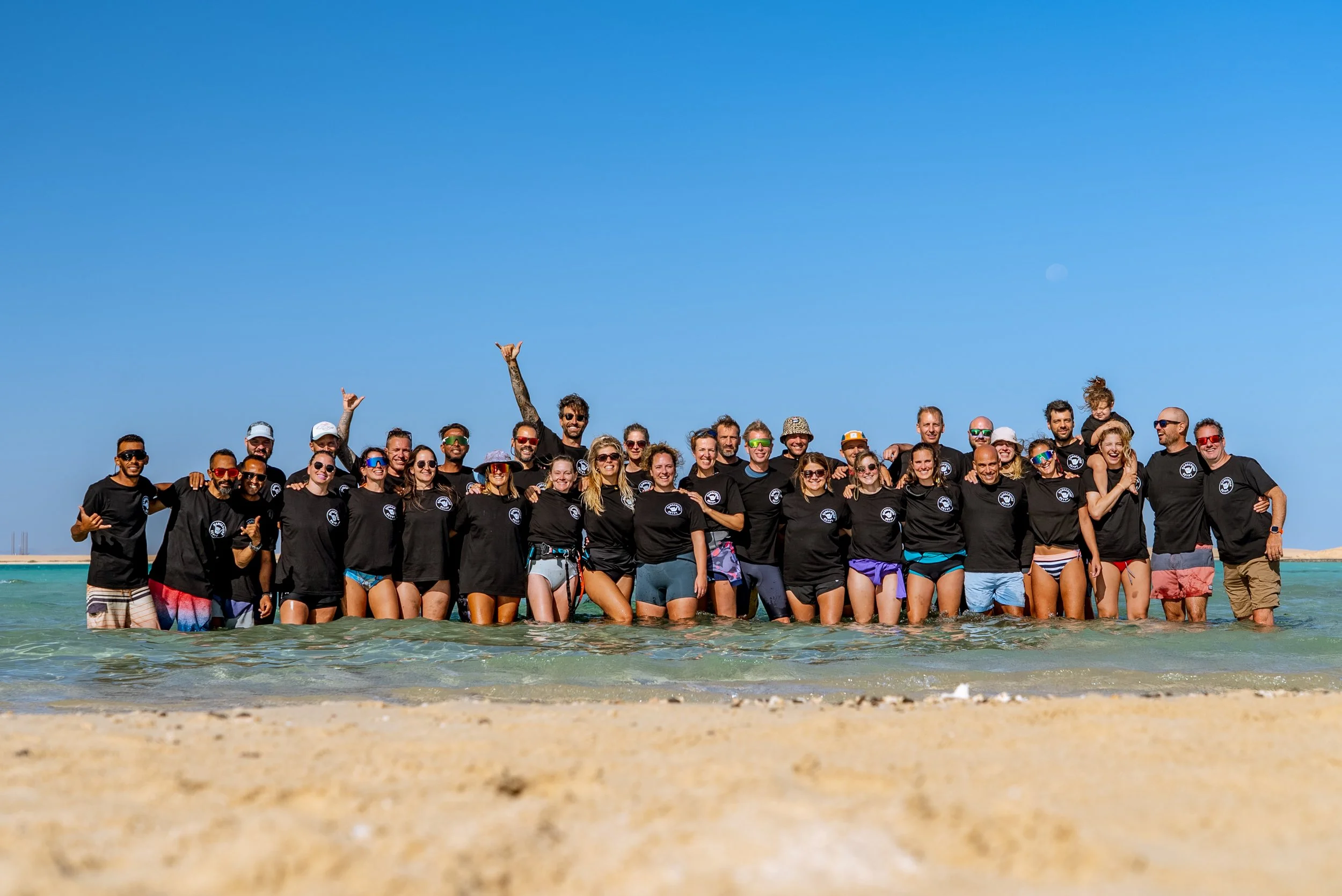 A large group of people standing in shallow ocean water on a beach, wearing matching black t-shirts, with clear blue sky in the background.