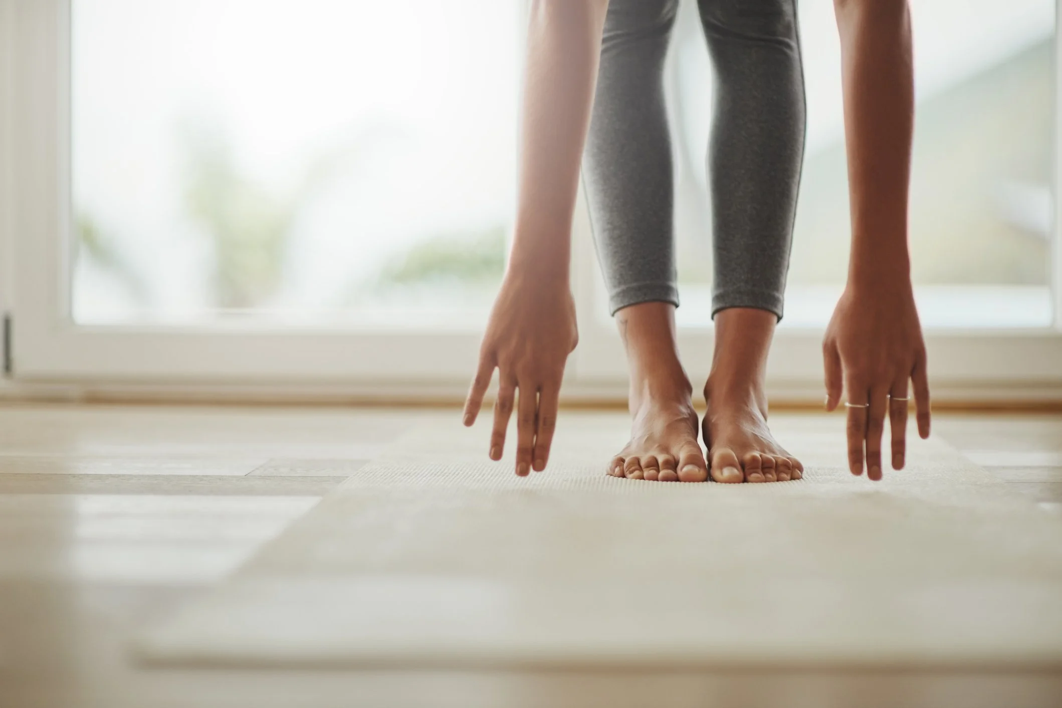 Person practicing yoga in a bent forward fold pose on a light-colored mat in front of a large window.