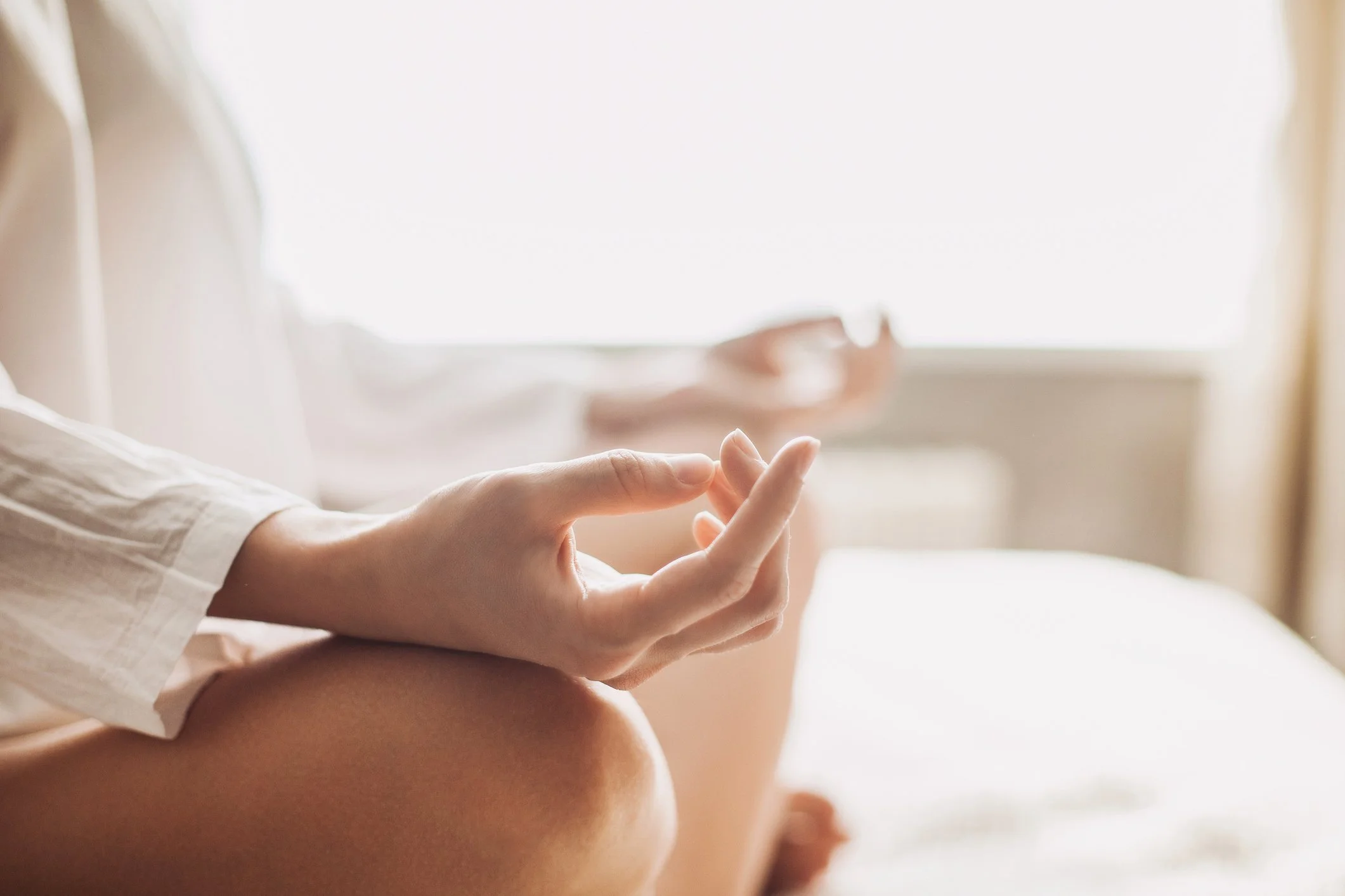 Close-up of a person sitting cross-legged and meditating with hands in a meditation mudra indoors near window with sunlight.