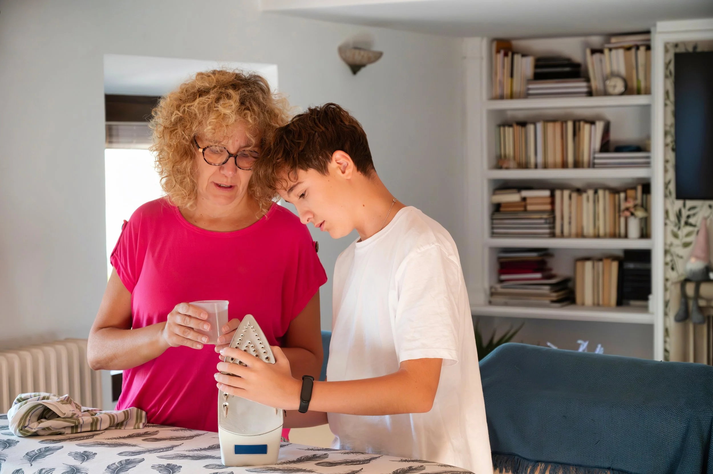 A woman with curly blonde hair and glasses, wearing a bright pink shirt, and a young boy with short brown hair, wearing a white t-shirt, are looking down at an iron placed on a table. The woman is holding a pink mug and the boy is holding the iron, both appearing focused.