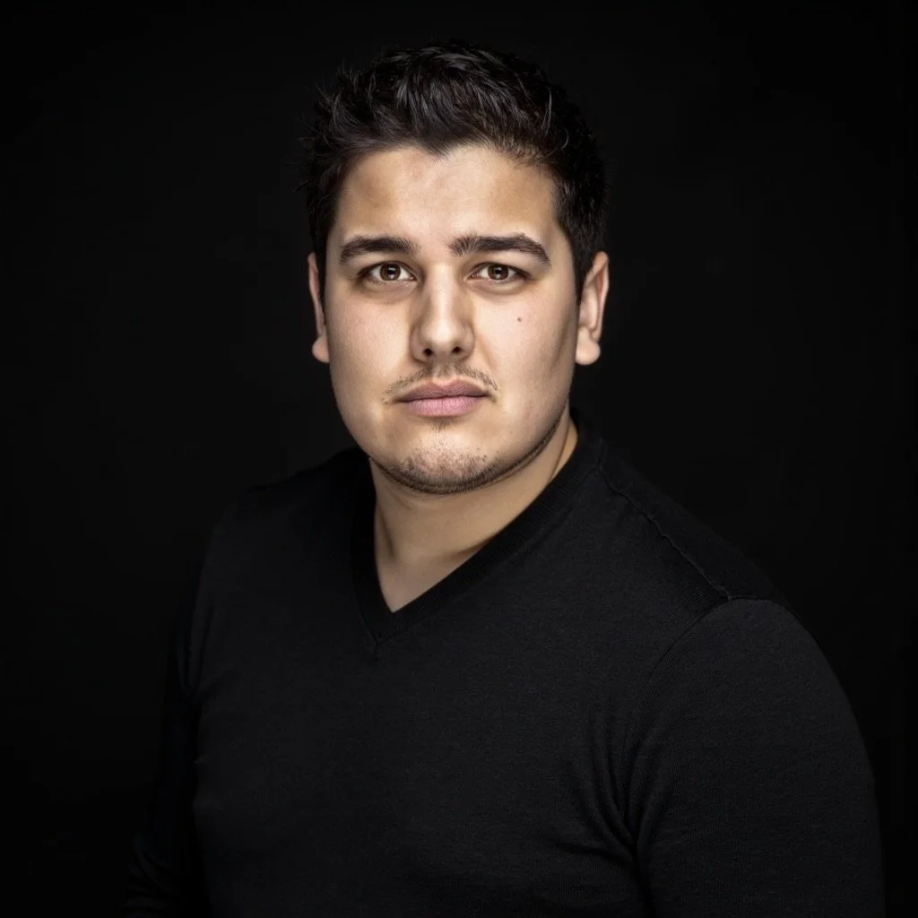 Alt text: Hamish Wyllie, a man with short dark hair and light stubble, wearing a black V-neck shirt, looks confidently at the camera against a dark background.