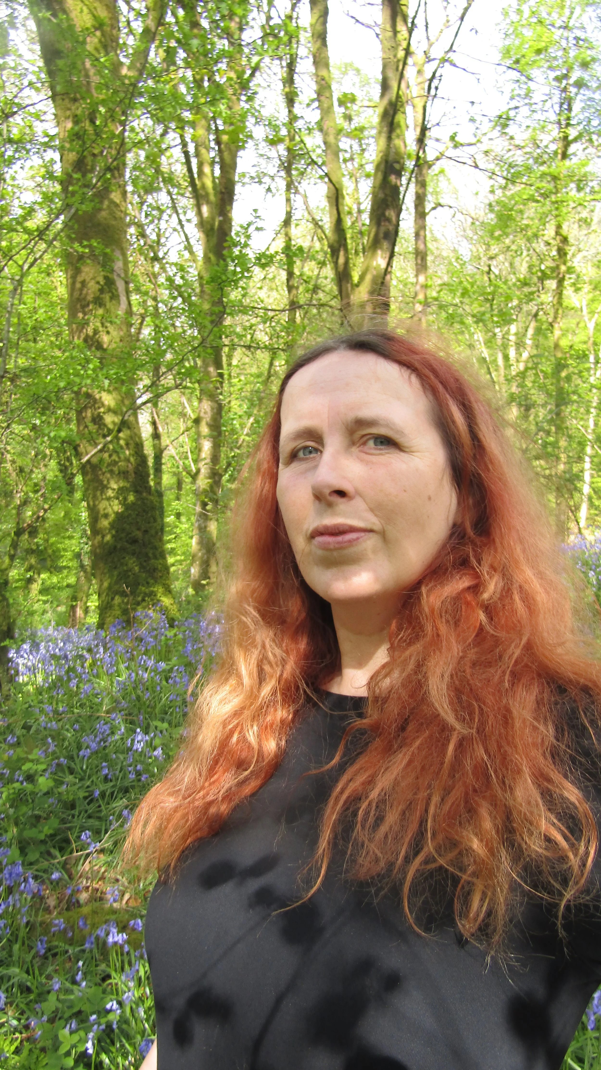 Alt text: Ruth Urquhart, a woman with long reddish-brown hair wearing a black top, stands outdoors in a sunlit forest surrounded by trees and blooming bluebells, looking calmly at the camera.