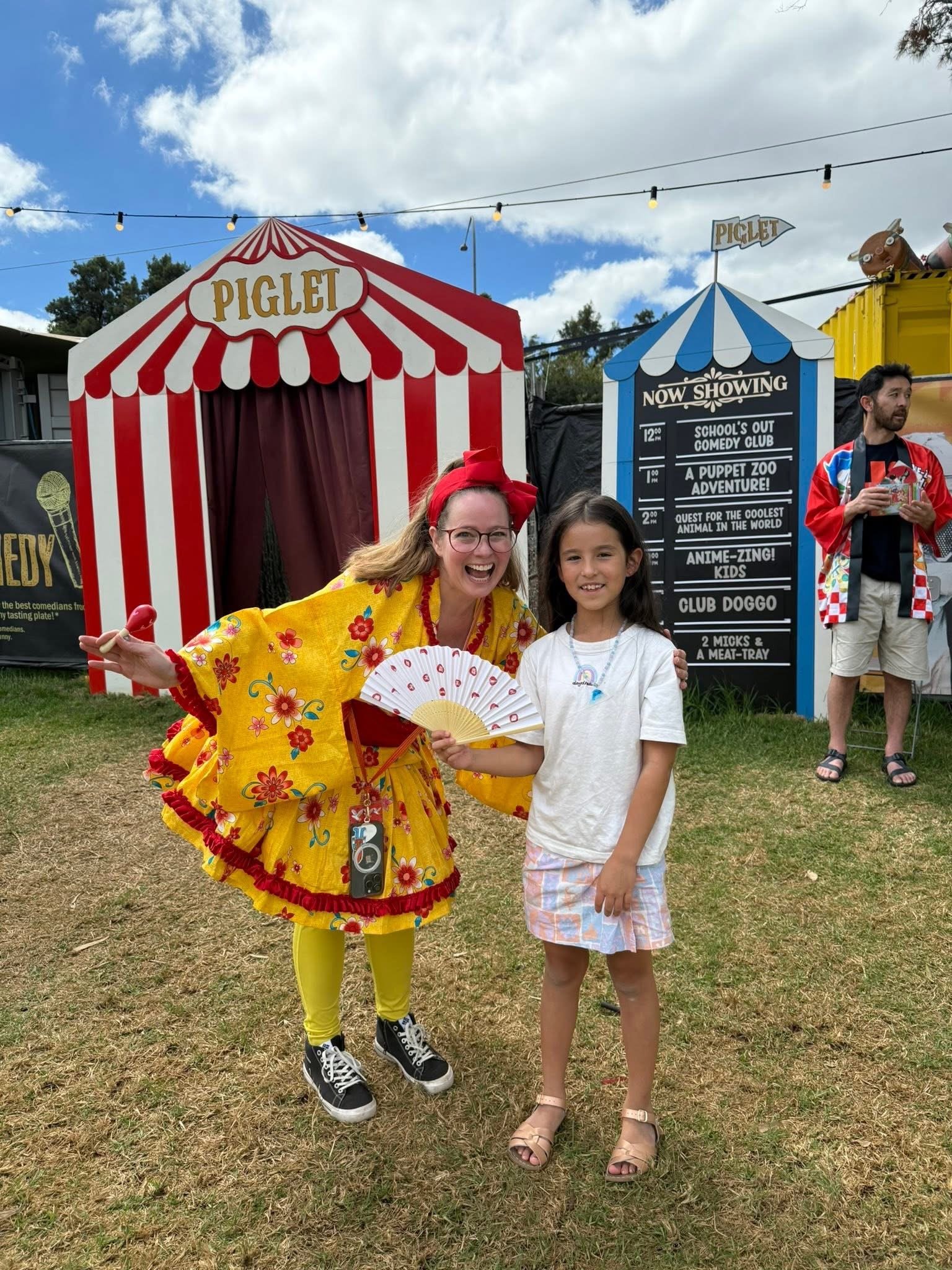 A woman dressed as a clown with yellow floral costume and yellow tights is standing next to a young girl wearing a white shirt and plaid shorts, at an outdoor fair with carnival booths and a cloudy sky in the background.