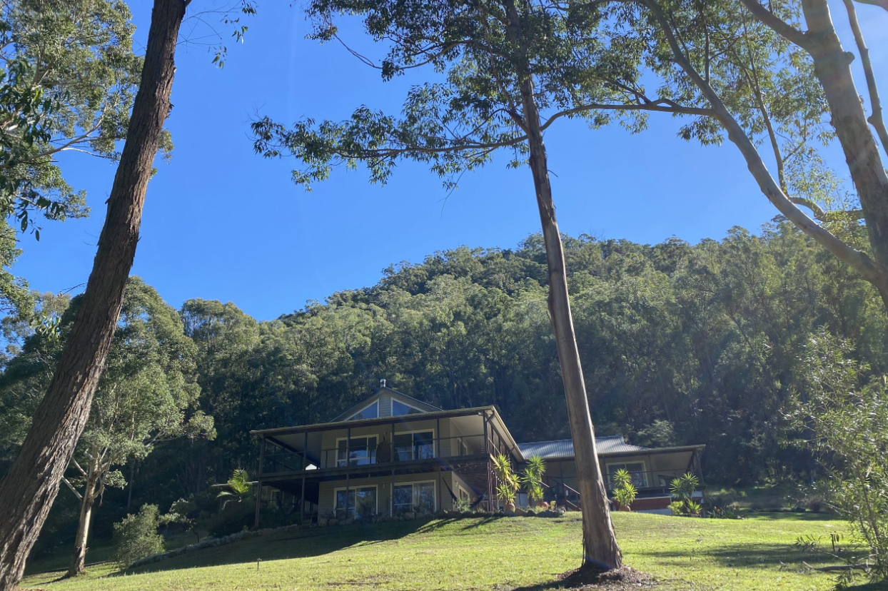 A house with large decks on multiple levels, set on a grassy slope in a wooded area with trees and a blue sky.