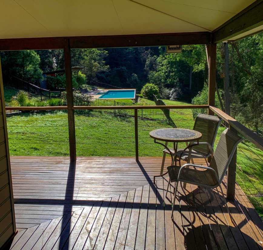 Wooden balcony with two chairs and a small round table overlooking a grassy yard and a pool in the distance, surrounded by trees.