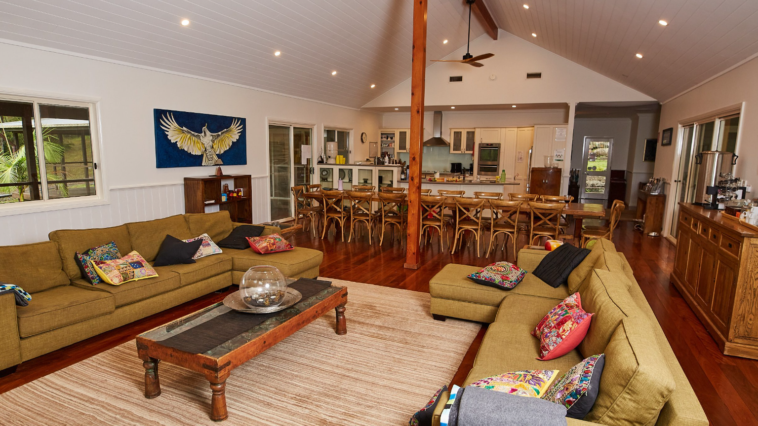 A spacious open-concept living and dining area with wooden flooring, white paneled walls, and a vaulted ceiling with recessed lighting. The living room has two olive-green sofas with colorful pillows, a wooden coffee table, and a fishbowl centerpiece