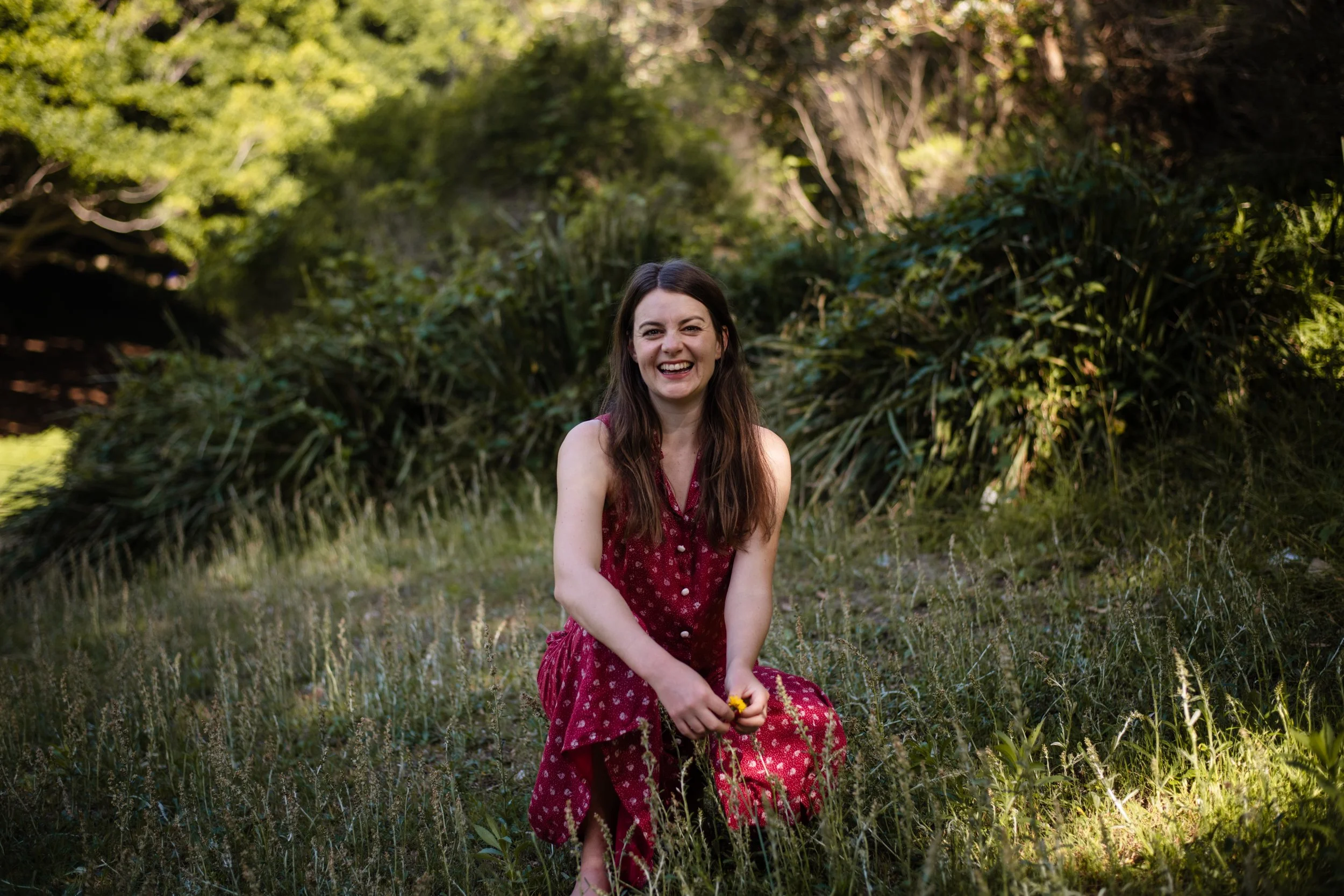 A woman with long brown hair smiling and sitting outdoors in a grassy area, wearing a red dress with white polka dots, holding a small yellow flower, surrounded by green trees and bushes.