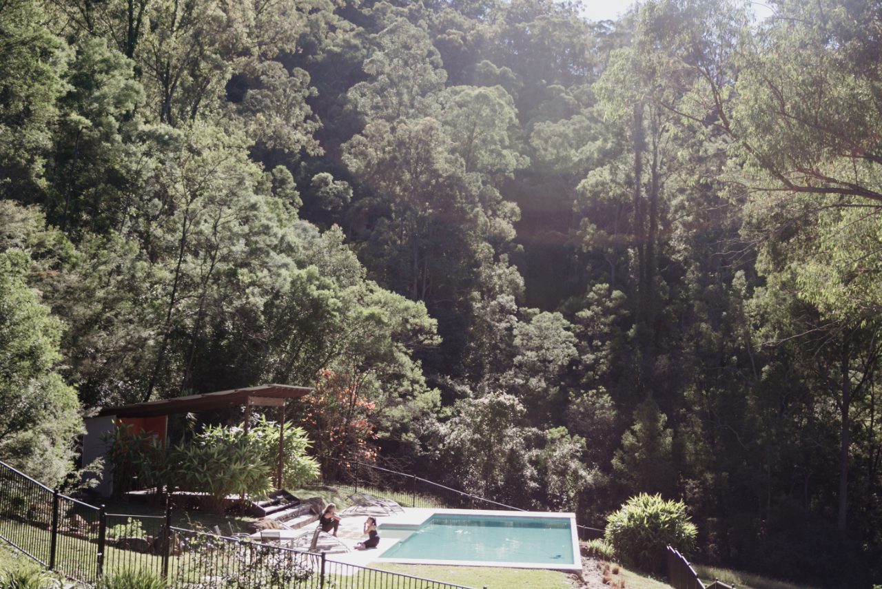 A backyard with a small pool, surrounded by trees and greenery on a hillside, with two people sitting near the pool.