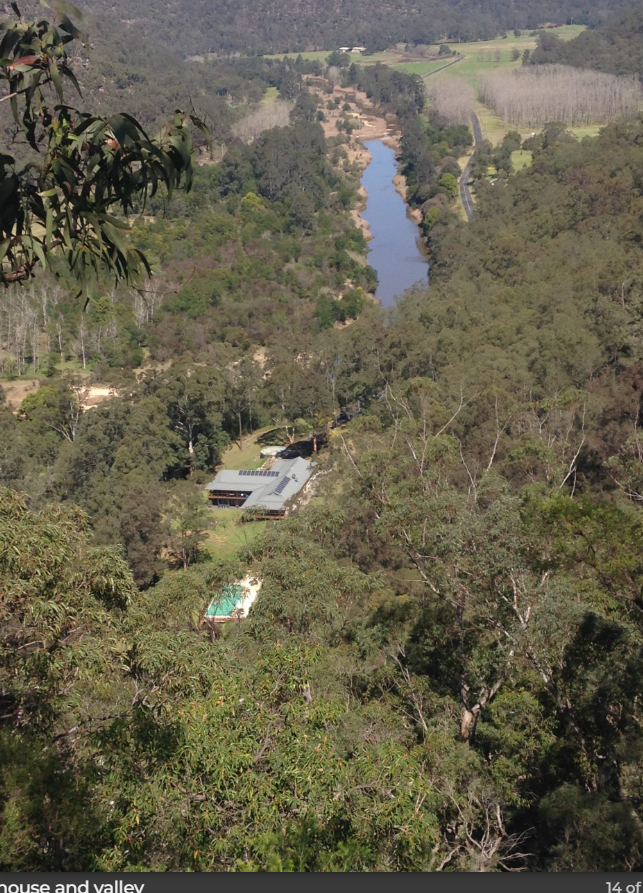 Aerial view of a house and valley with a river running through the middle, surrounded by dense trees and open green fields.