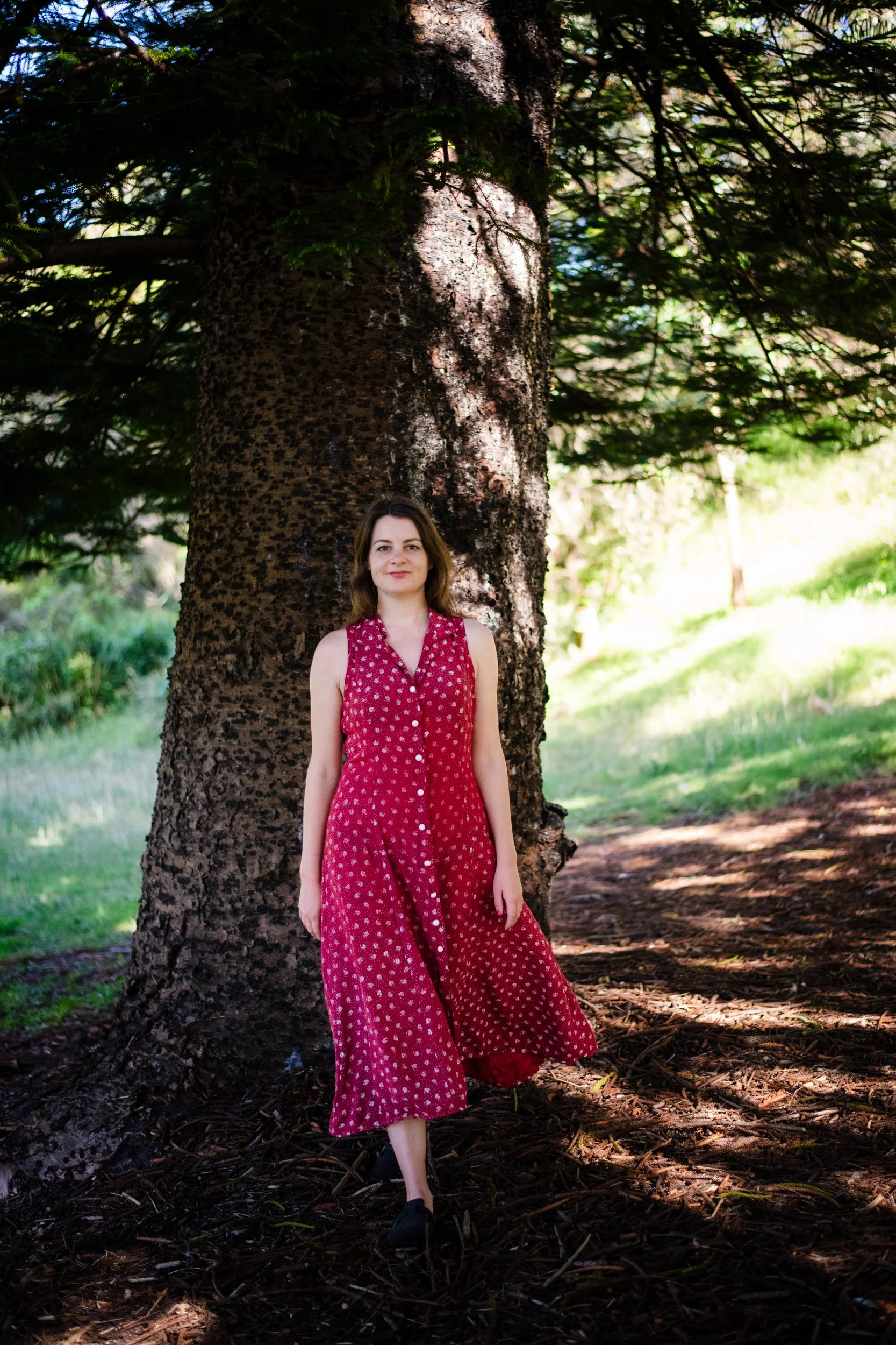 A woman in a red dress with small white polka dots standing in front of a large tree in a forested area