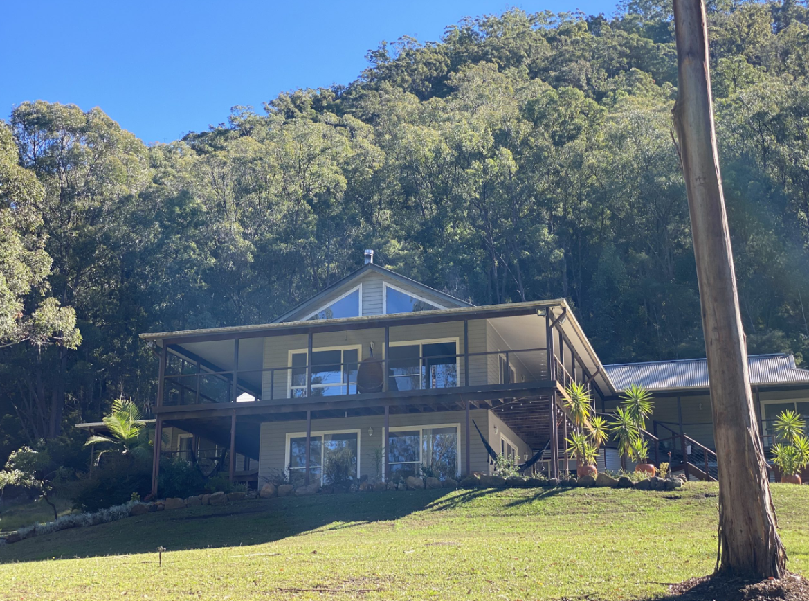 A two-story house with a screened-in porch on the lower level and a balcony on the upper level, situated on a grassy hill with trees and a mountain in the background under a clear blue sky.