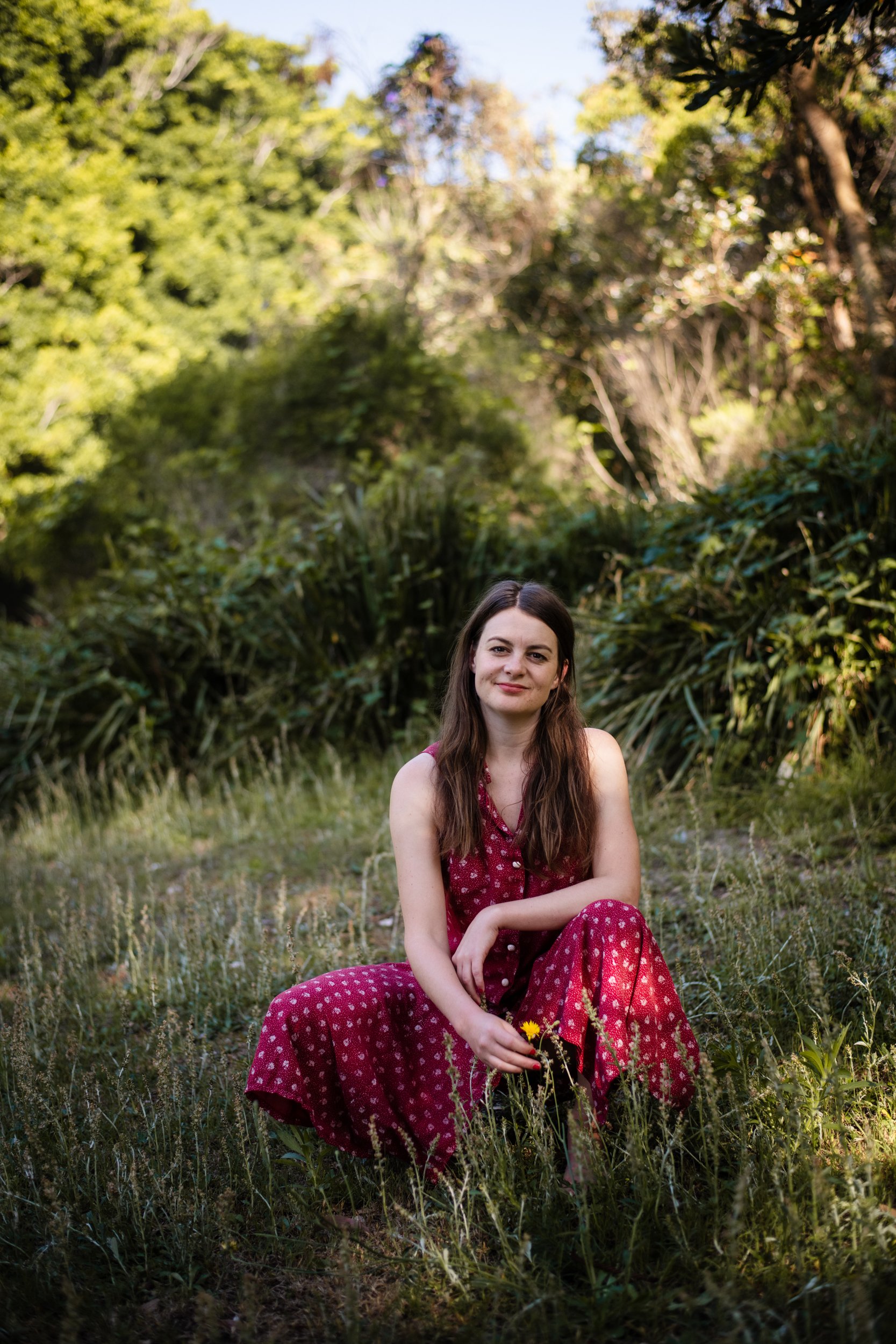 A young woman with long brown hair, wearing a red polka dot dress, sitting on the grass in a lush outdoor setting with trees and greenery in the background, holding a small yellow flower.