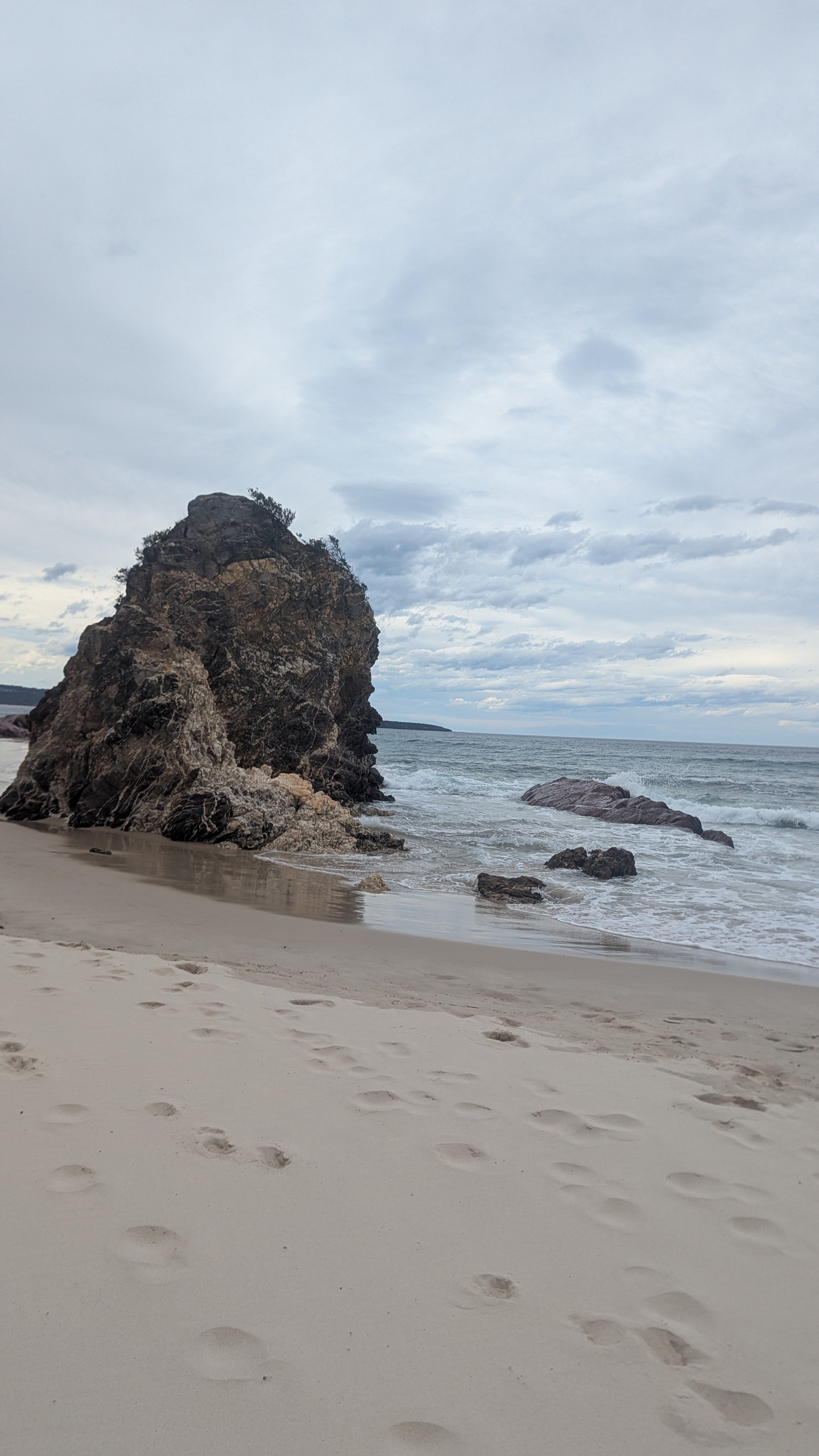 A beach scene with footprints in the sand, large rocks near the shore, and the ocean extending to the horizon under a cloudy sky.