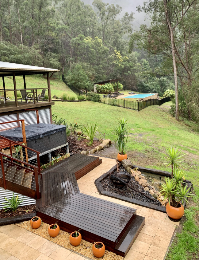 View of a backyard with a wooden deck, potted plants, a hot tub, and a fenced swimming pool area surrounded by trees.