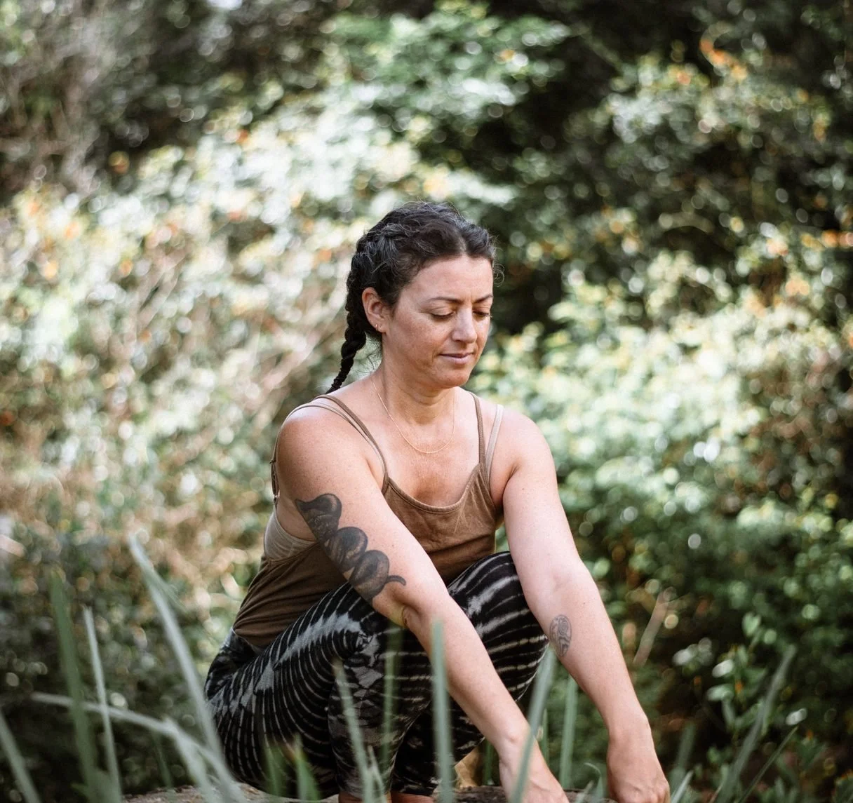 A woman with braided dark hair, tattoos on her arms, wearing a brown tank top and striped pants, crouches outdoors in a forested area with greenery.