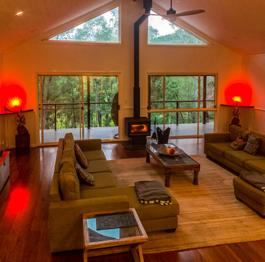 Living room with vaulted ceiling, large triangular windows, and sliding glass doors leading to a balcony overlooking trees. There is a central wood-burning stove with a chimney, two green sofas with colorful pillows, a wooden coffee table, a mix of w