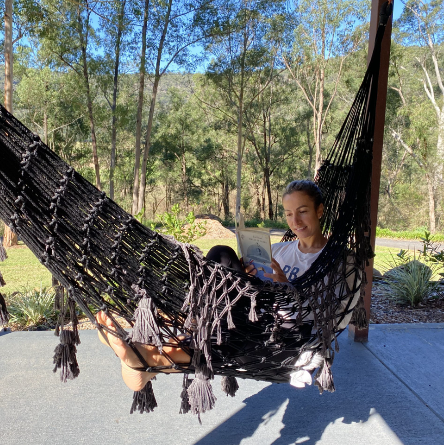 A woman relaxing in a black hammock outdoors, reading a book, with trees and a clear sky in the background.