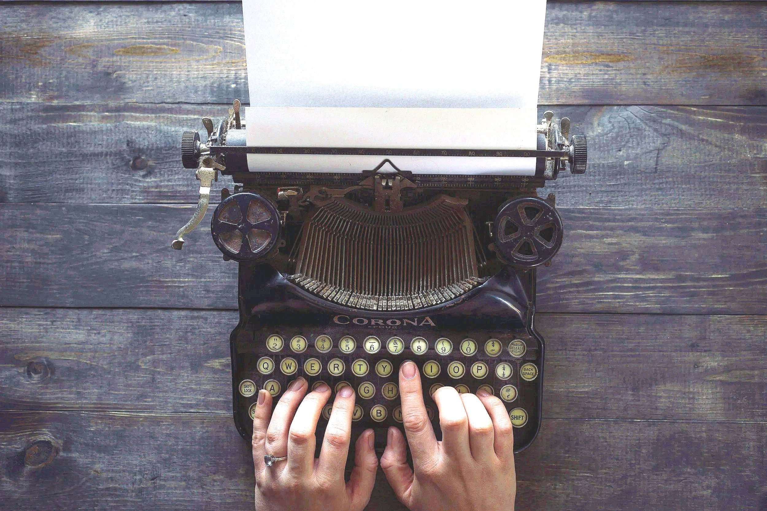 Persoon typt op een vintage typemachine van het merk Corona met een stuk papier erin, op een donkere houten tafel.