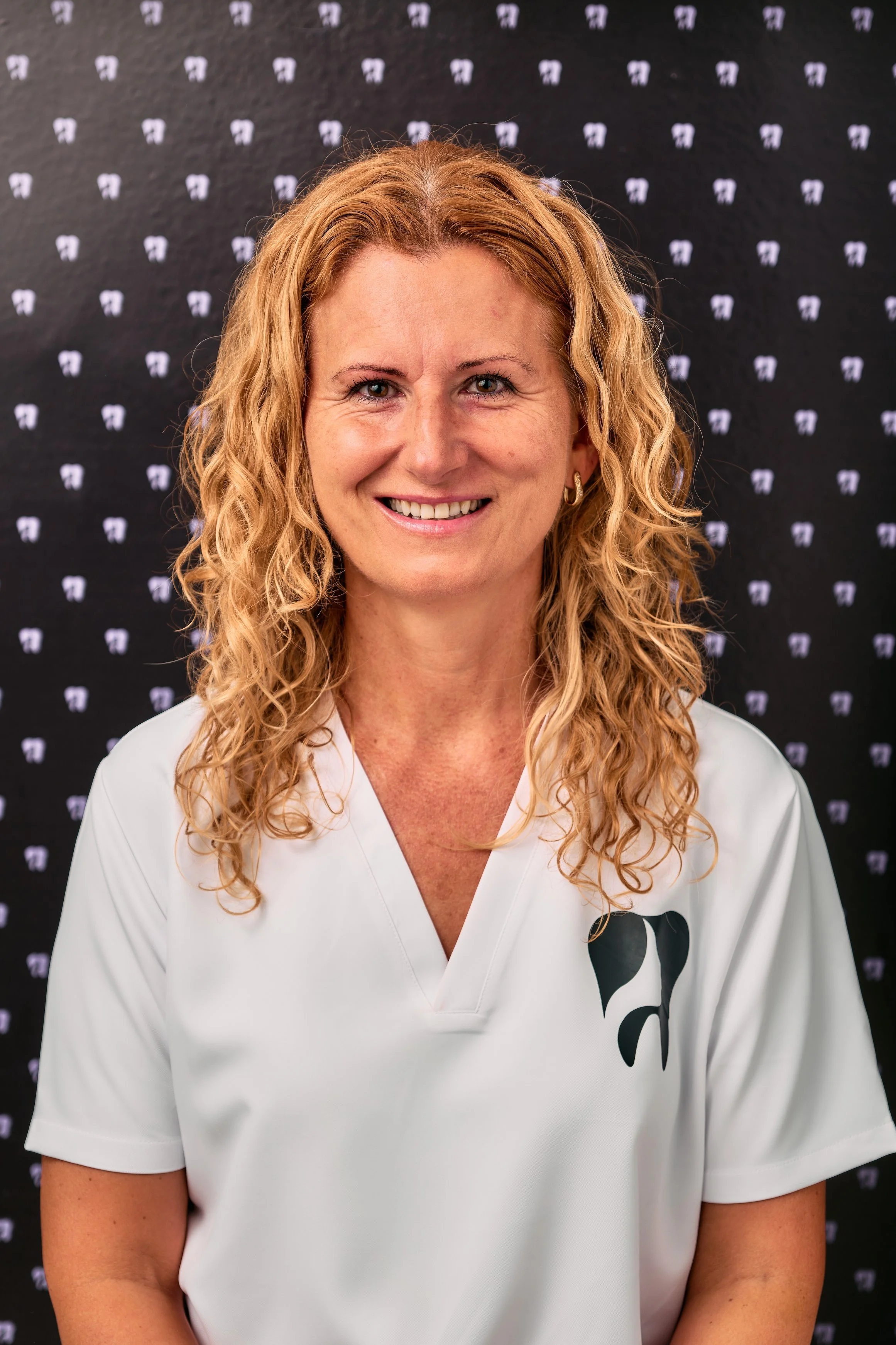 A woman with curly red hair wearing a white top with a black logo, smiling at the camera, standing in front of a black backdrop with small white and purple design elements.