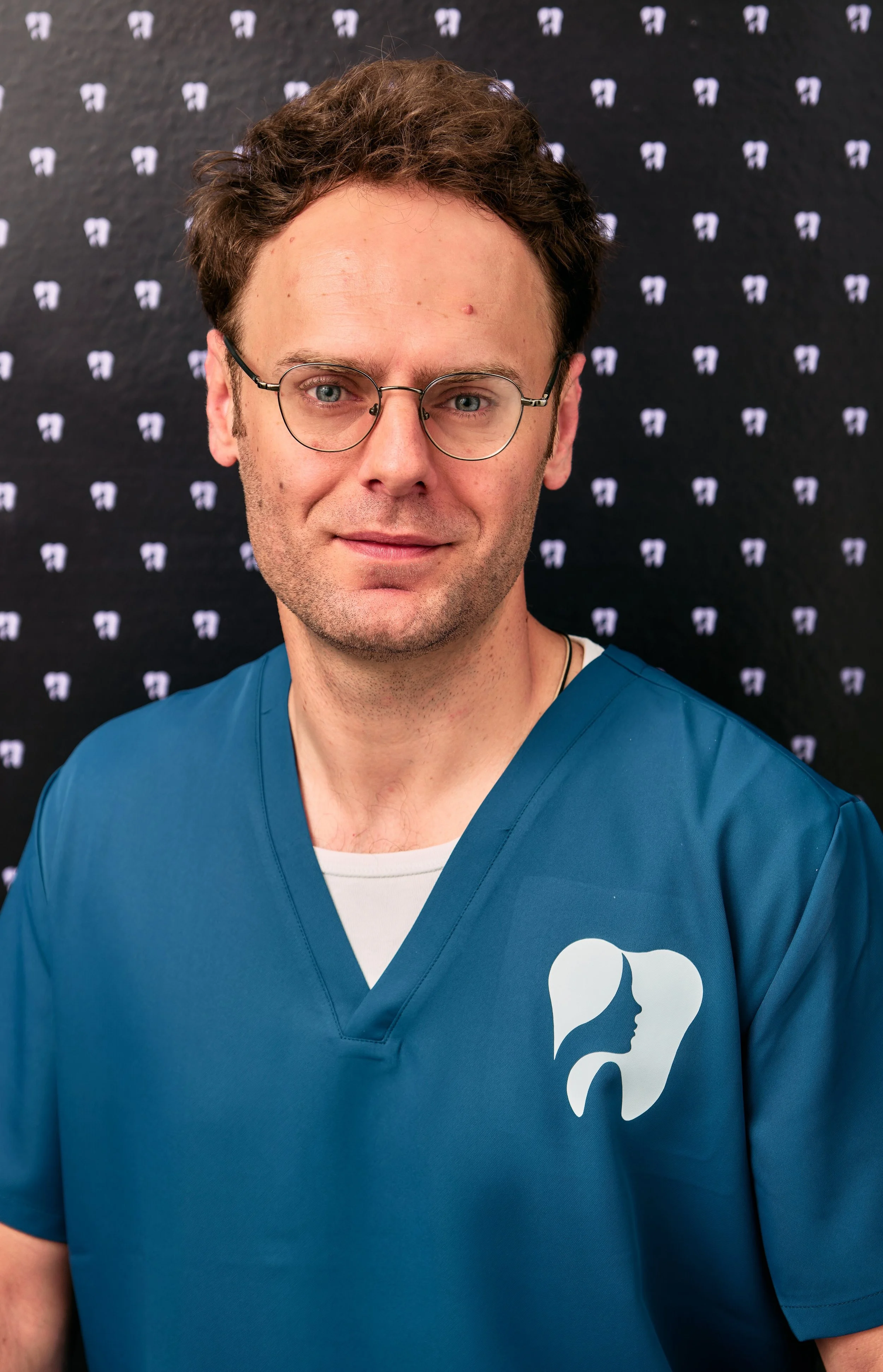 A man with brown hair, glasses, and light skin, wearing a blue medical scrub top with a logo featuring a profile of a woman's face with long hair, standing in front of a black background with small white logos.