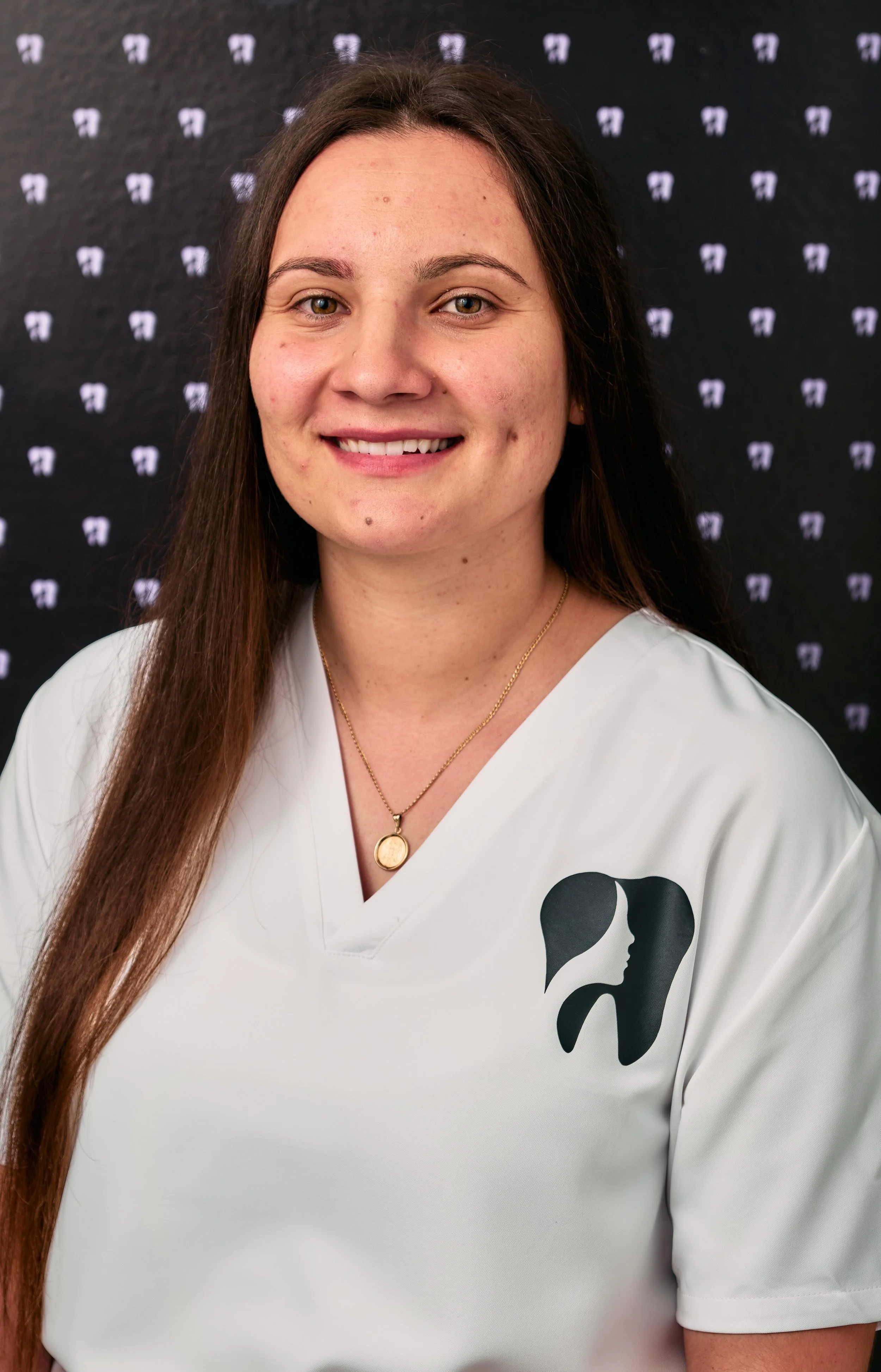 A woman wearing a white V-neck shirt with a black logo of a face and hair, standing against a dark background with small white symbols, smiling at the camera.