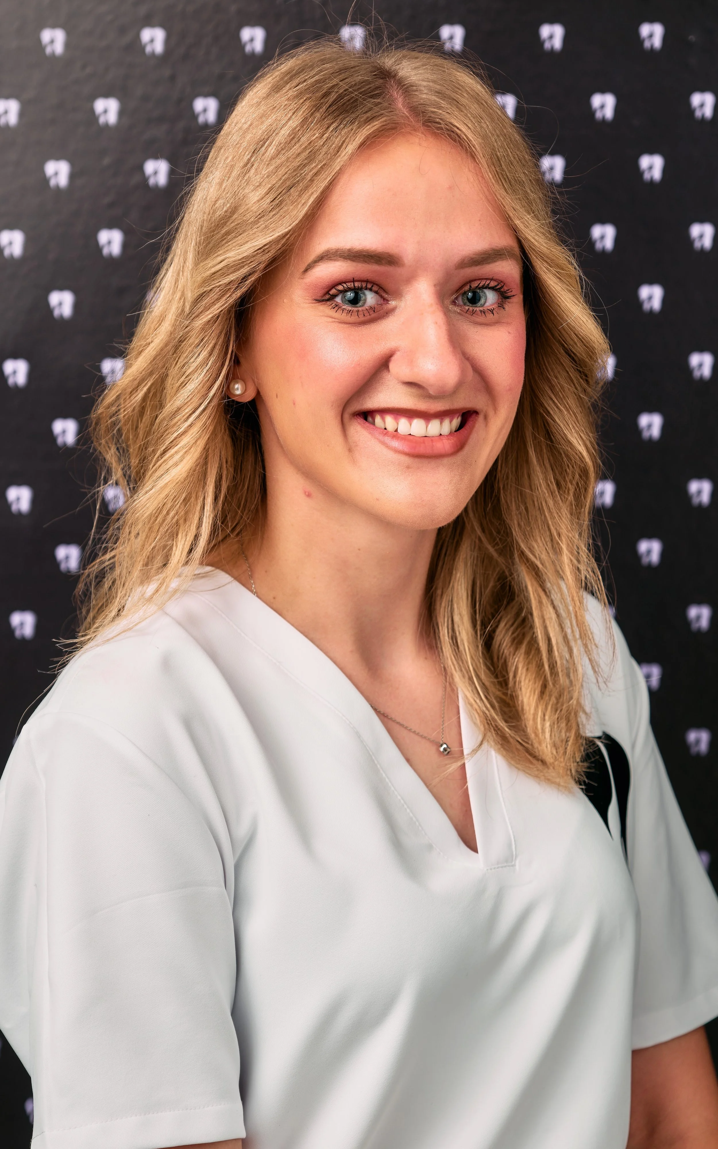 Young woman with blonde hair, blue eyes, and a friendly smile, wearing a white medical uniform, standing in front of a dark backdrop with a repeated white logo.