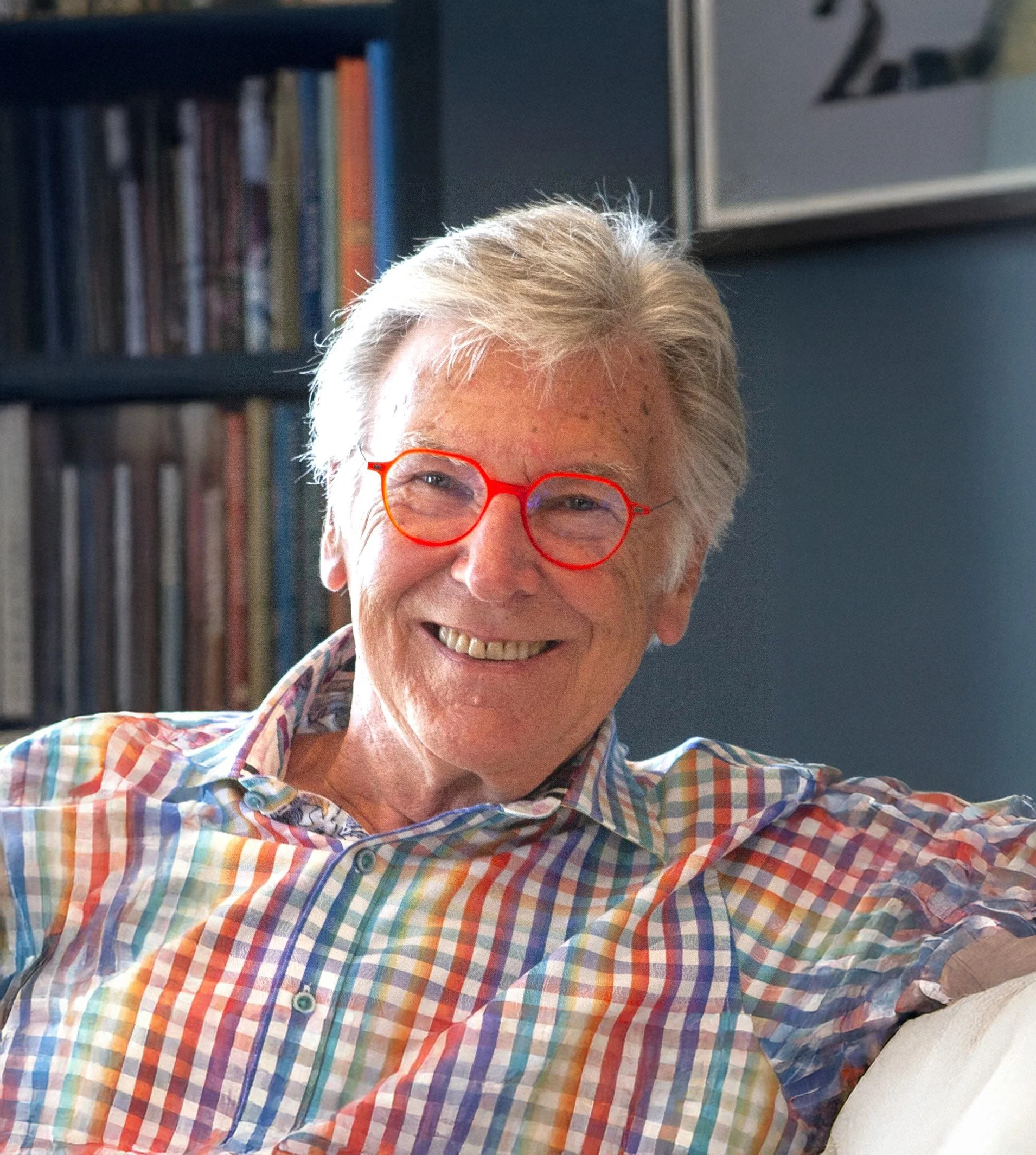 A smiling elderly man with grey hair, wearing bright red glasses and a colorful plaid shirt, sitting on a couch with a background of bookshelves and artwork.