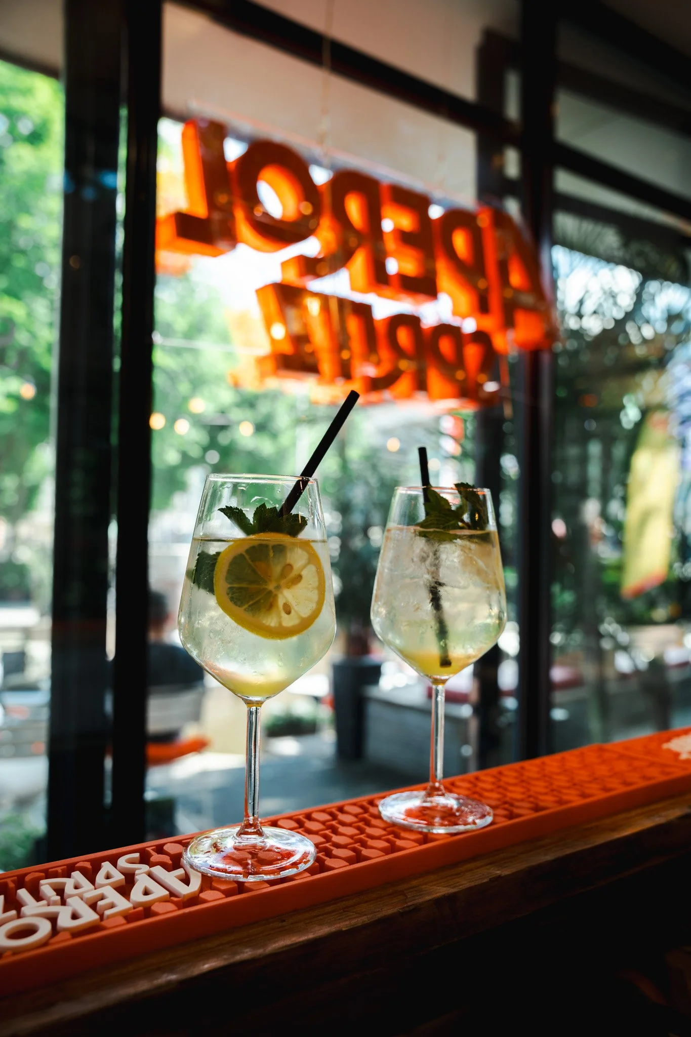 Two glasses of lemonade garnished with lemon slices and mint leaves on a bar counter, with a glowing orange 'OPEN' sign in the background at a sunny outdoor cafe.